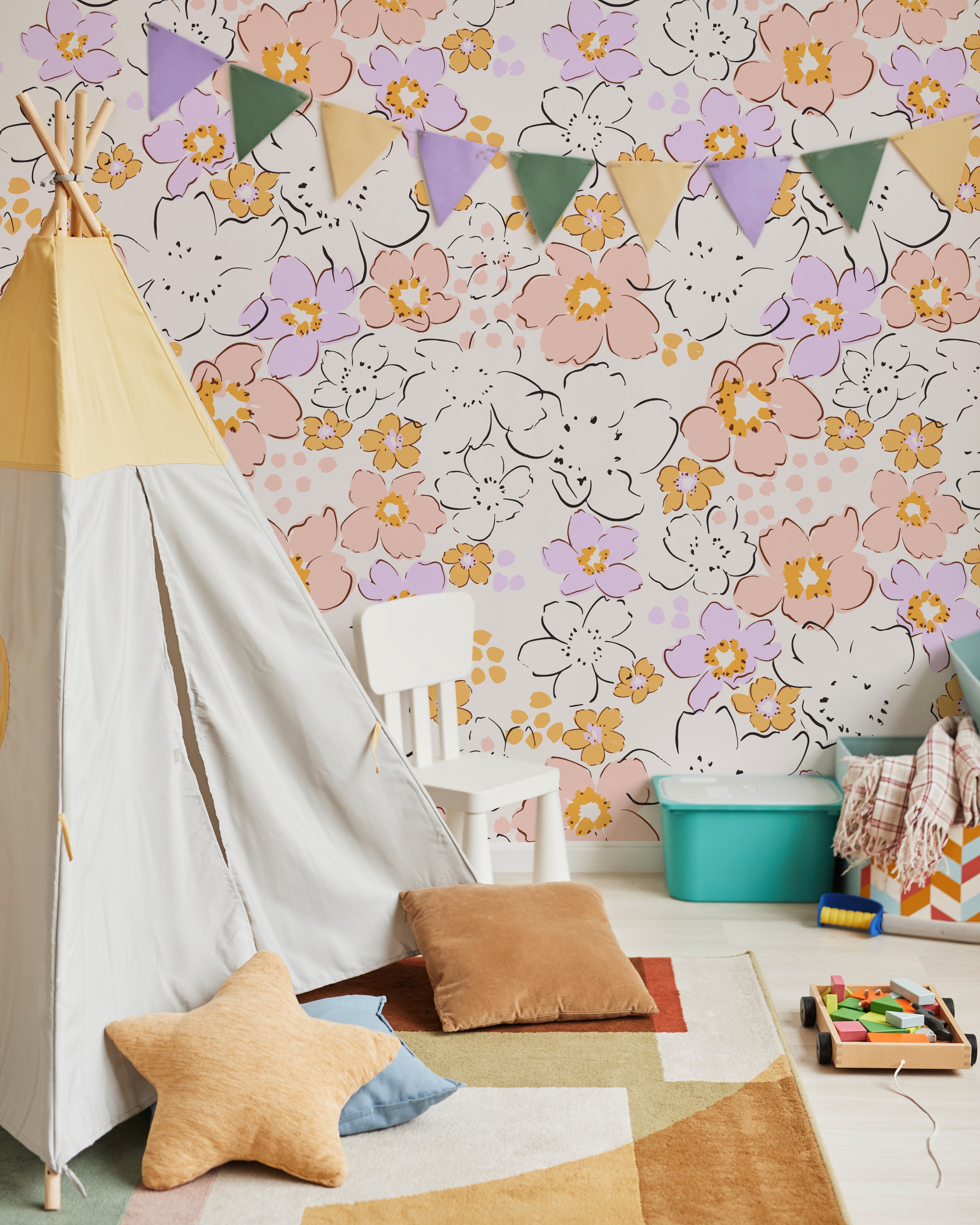 A vibrant and playful children's play area with a wall adorned by the Ditsy Garden Wallpaper, featuring a cheerful pattern of white, pink, and yellow flowers on a soft pastel background. The scene includes a white teepee, colorful hanging pennants, and a variety of toys and cushions, creating a joyful and inviting space.