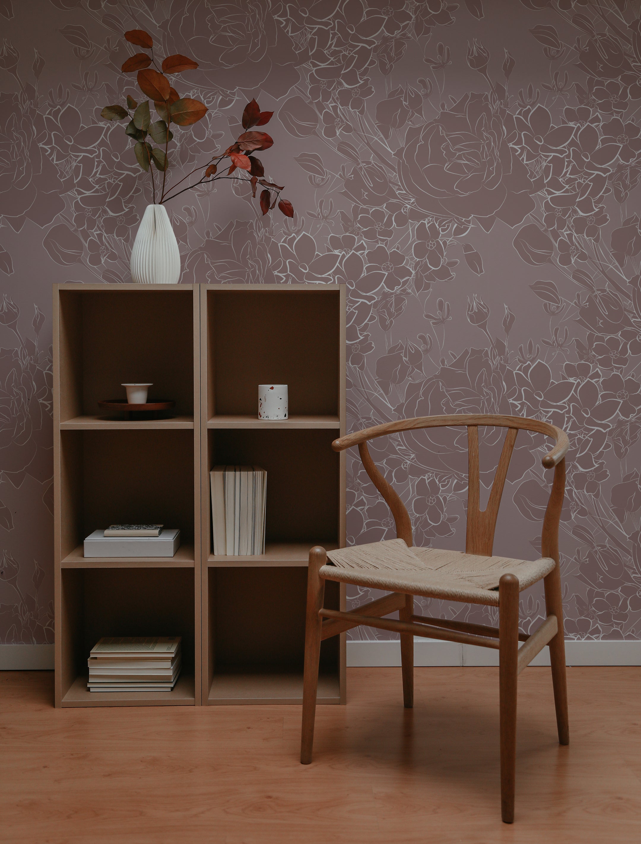 A cozy reading nook with Dusty Rose Floral Wallpaper adorning the wall, complemented by a simple wooden chair and a shelving unit filled with books. The soft pink background and white floral patterns evoke a serene, inviting space