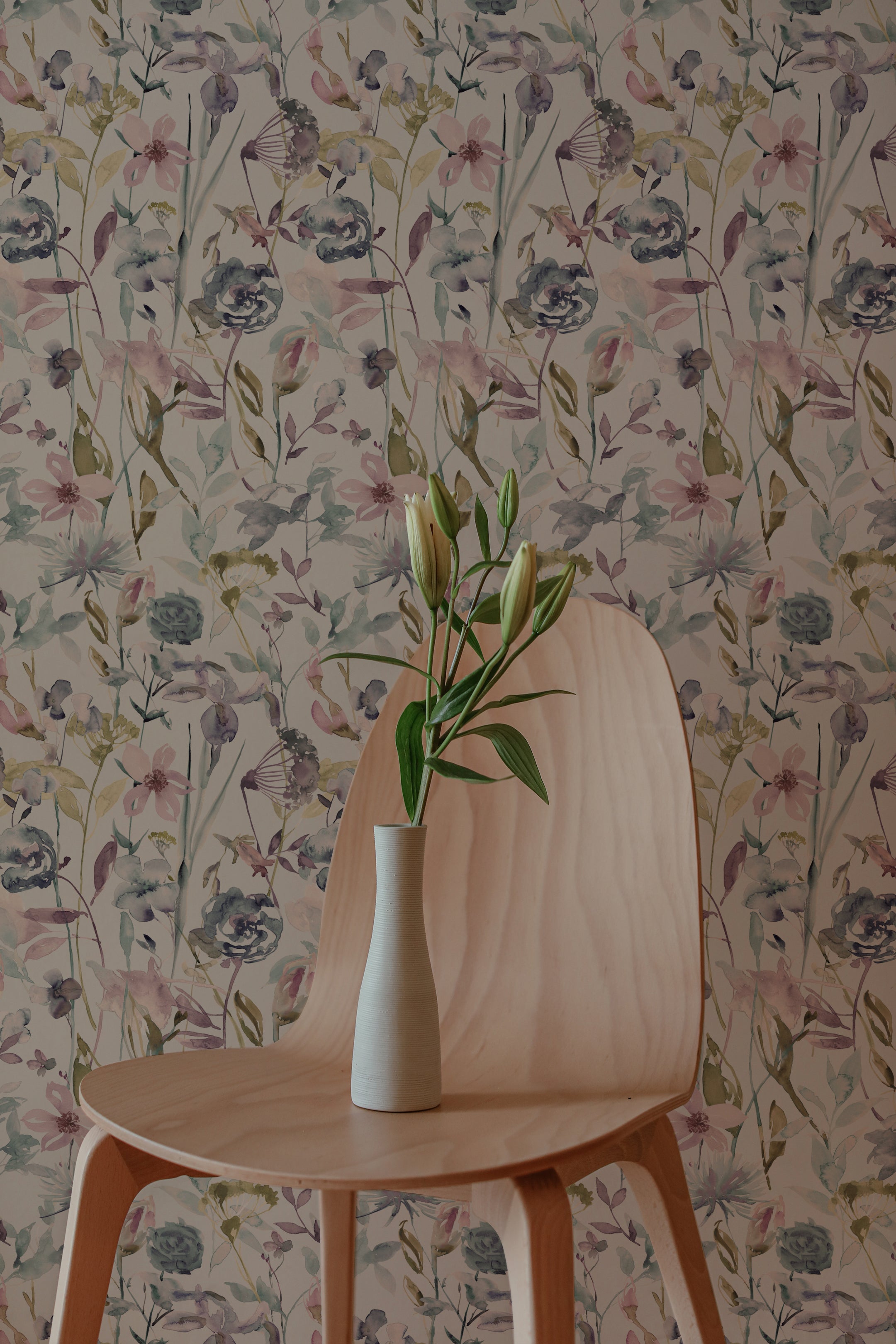 A minimalistic interior featuring a modern chair with a vase of fresh lilies on it. The background showcases the Watercolour Meadow Wallpaper, with its soft watercolor floral pattern in shades of pink, blue, and green, giving a serene and artistic touch to the space.