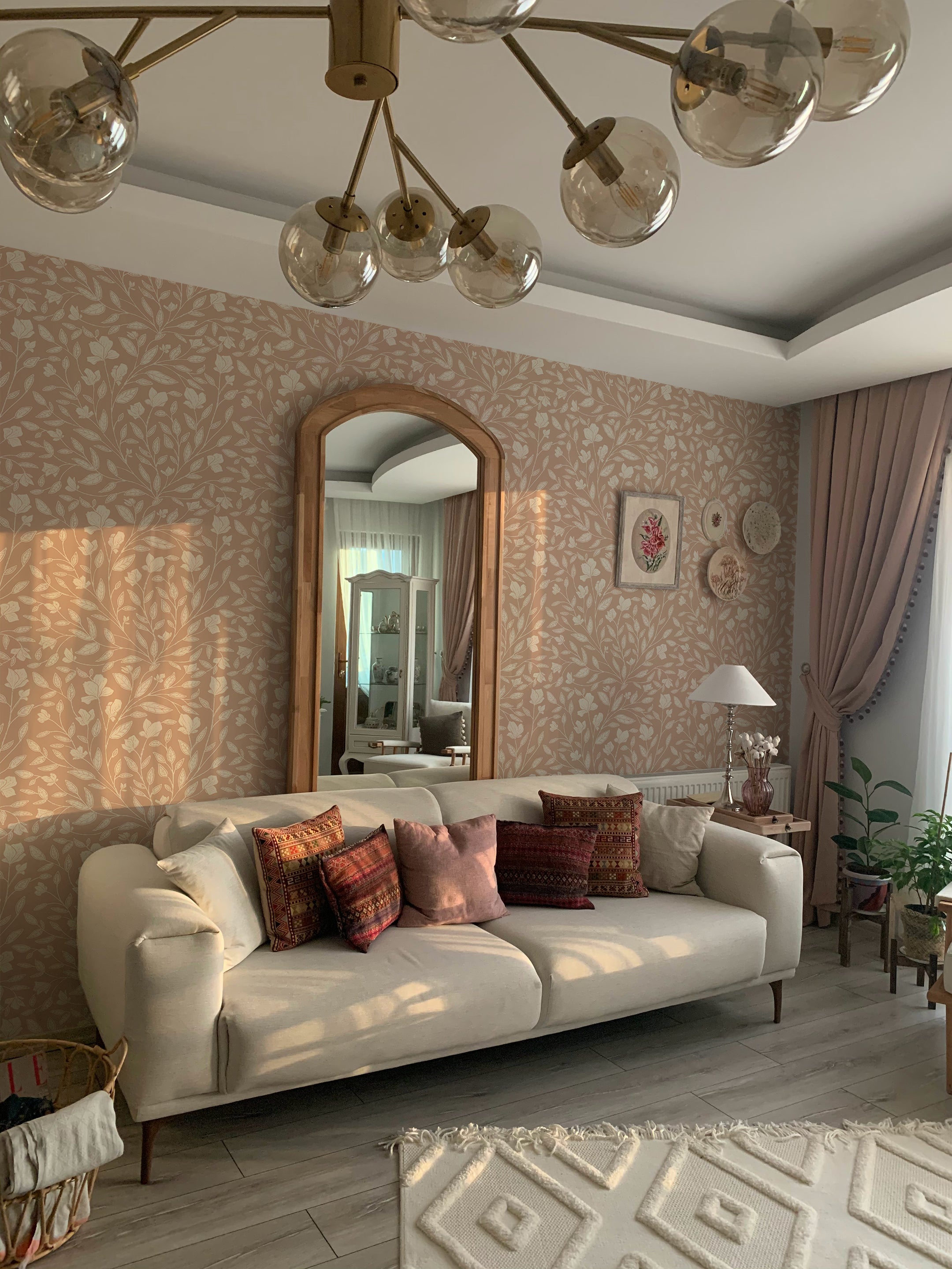 An elegantly furnished living room featuring a taupe sofa with vibrant patterned cushions, illuminated by a modern chandelier, against a backdrop of taupe floral vine wallpaper. The room is accented with a large arched mirror and decorative plates, creating a cozy and stylish atmosphere.