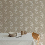 A dining setup featuring a simple white tablecloth and minimalist tableware on a rustic wooden table, set against a backdrop of a wall covered in a floral chintz wallpaper with white blossoms on a linen background, creating a serene dining atmosphere
