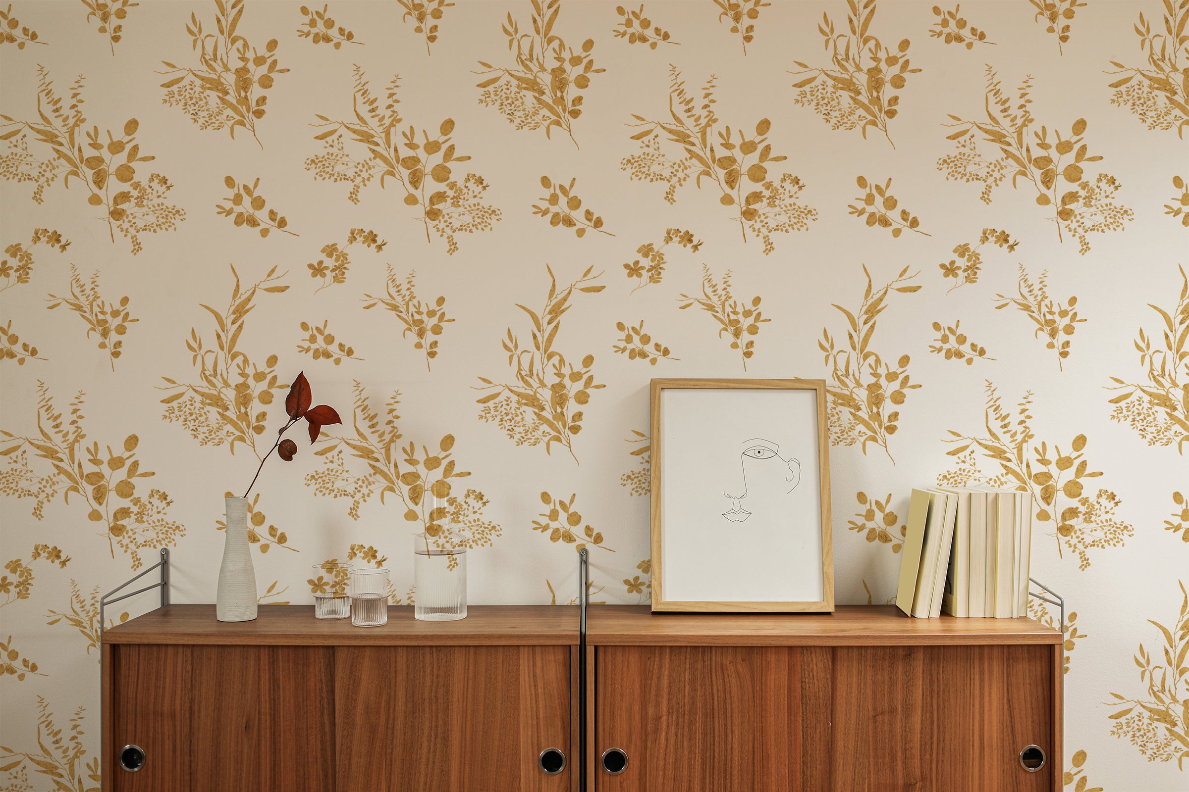 A minimalist decor arrangement against the Golden Greenery Wallpaper. A simple, modern wooden cabinet under the wallpaper is adorned with a small, elegant vase with a single dried plant, books, and glass containers, emphasizing simplicity and natural tones.