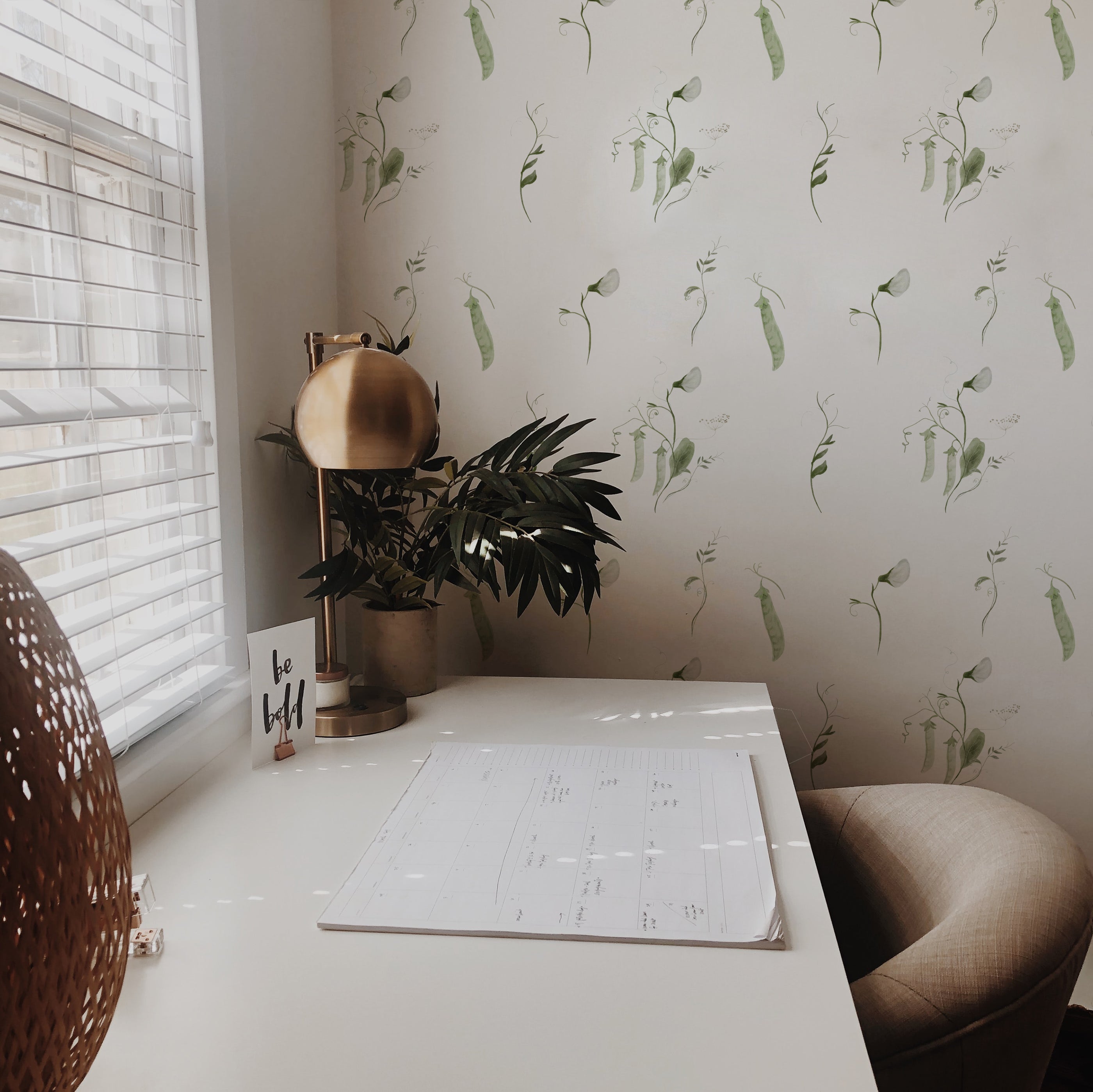 A cozy workspace corner illuminated by natural light filtering through a window with blinds. The room features a distinctive wallpaper decorated with delicate snow pea illustrations in soft green tones. The desk holds a calendar, a brass lamp with a leafy plant, and a motivational card reading "be bold."