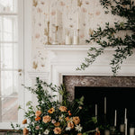 An elegant living space with Ikebana Floral Wallpaper, depicting soft-colored florals against a white background, alongside a classic white fireplace adorned with fresh greenery and tall candles, invoking a sense of sophisticated tranquility.