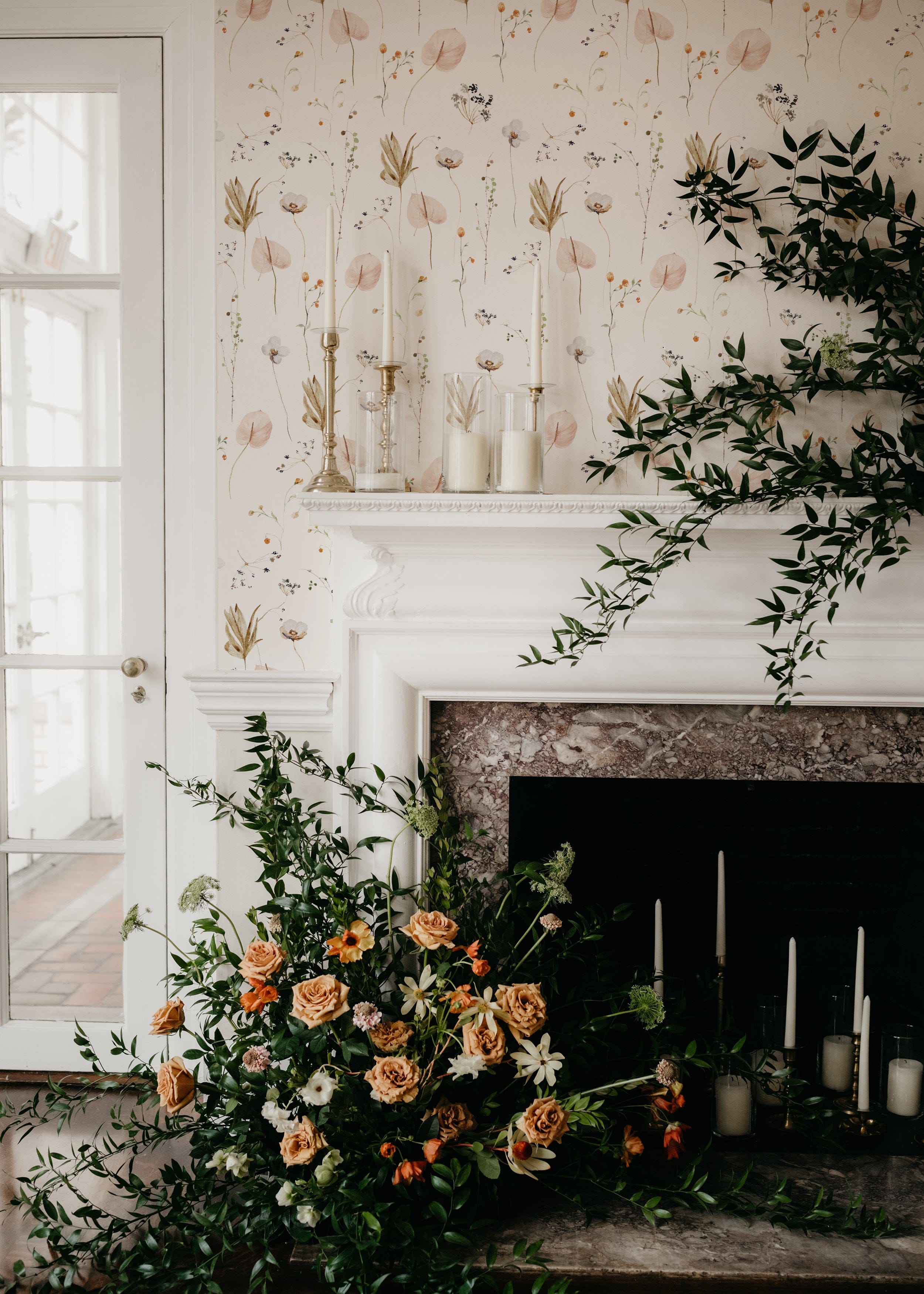 An elegant living space with Ikebana Floral Wallpaper, depicting soft-colored florals against a white background, alongside a classic white fireplace adorned with fresh greenery and tall candles, invoking a sense of sophisticated tranquility.