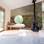 A contemporary room with a vintage wall mural depicting a rustic landscape. A wood-burning stove is positioned near the center, with a wooden bench and large circular mirror on the side. The scene blends seamlessly into a serene, forested backdrop visible through large windows.