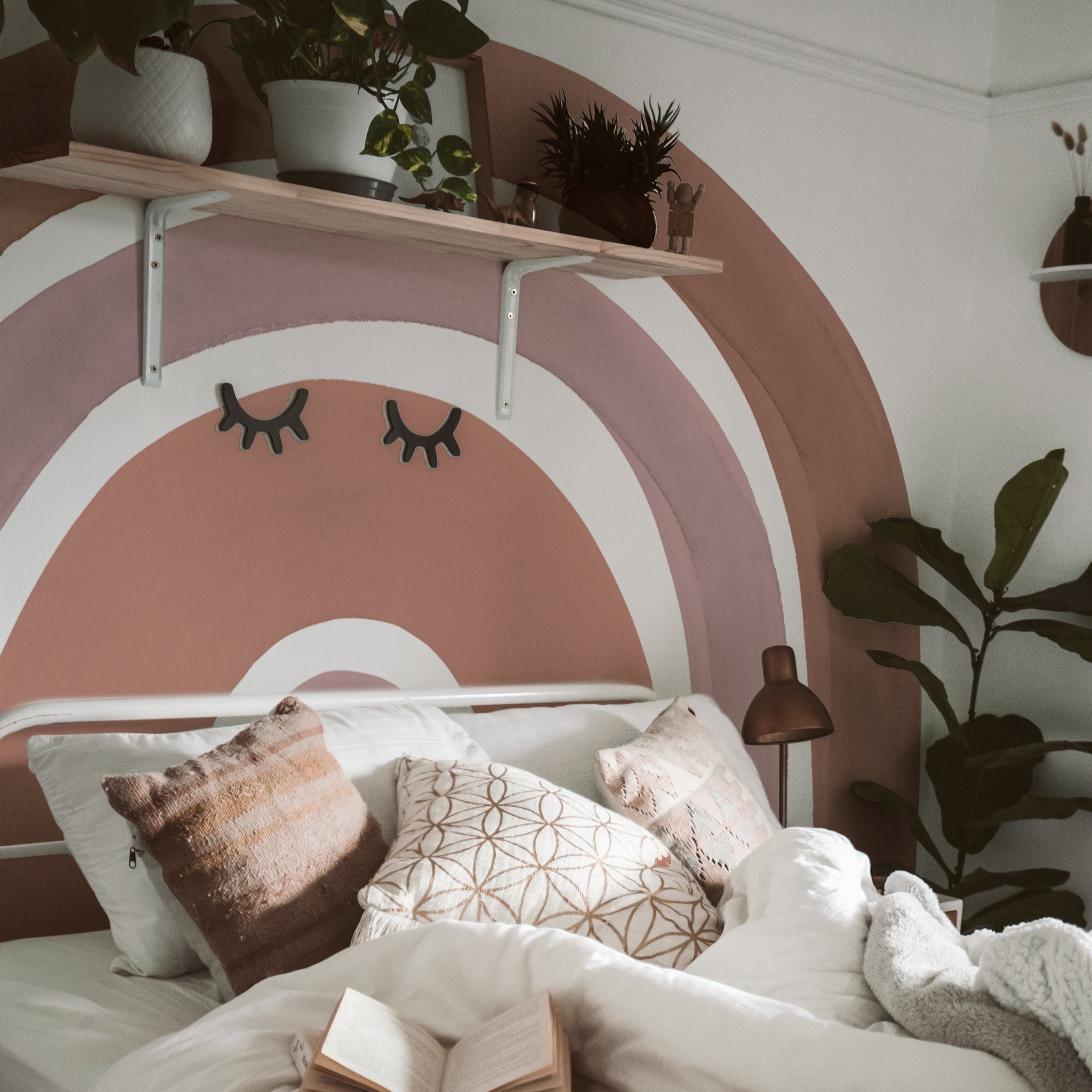 A cozy bedroom setting showcasing the Aesthetic Rainbow Wallpaper Mural - Luna. The mural features large, soft arches in muted tones of brown and pink, providing a warm backdrop. Decorated with plants and cozy bedding, this space reflects a tranquil and stylish ambiance.