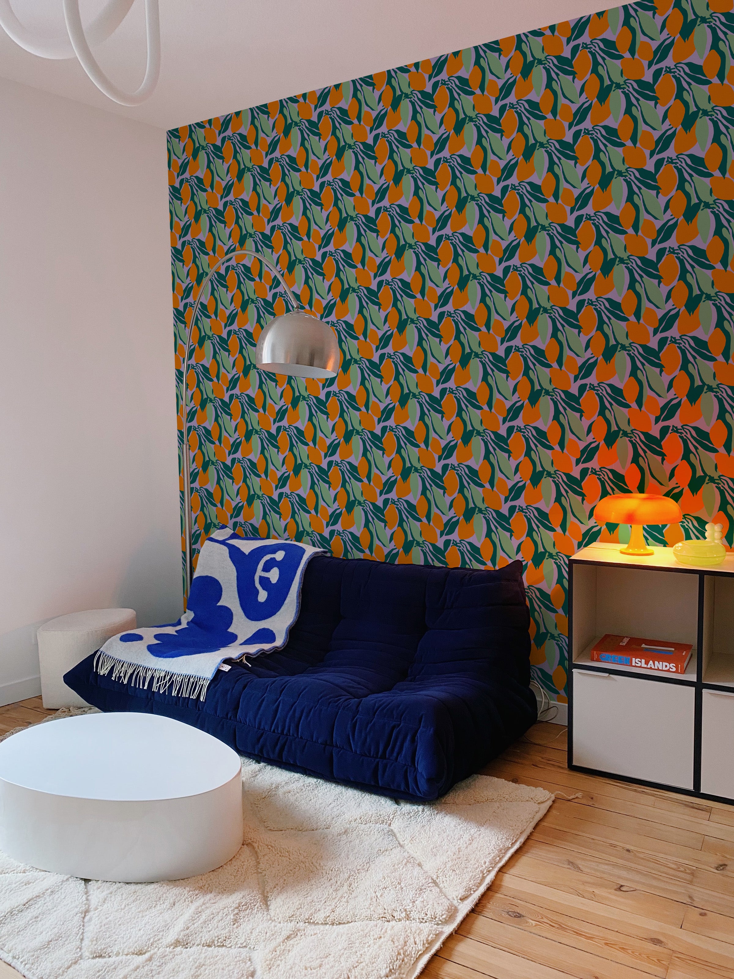 A modern living room featuring a wall covered in wallpaper with a pattern of orange fruits and green leaves on a purple background. The room includes a navy blue sofa, a round white coffee table, and various modern decorations, creating a vibrant and stylish space