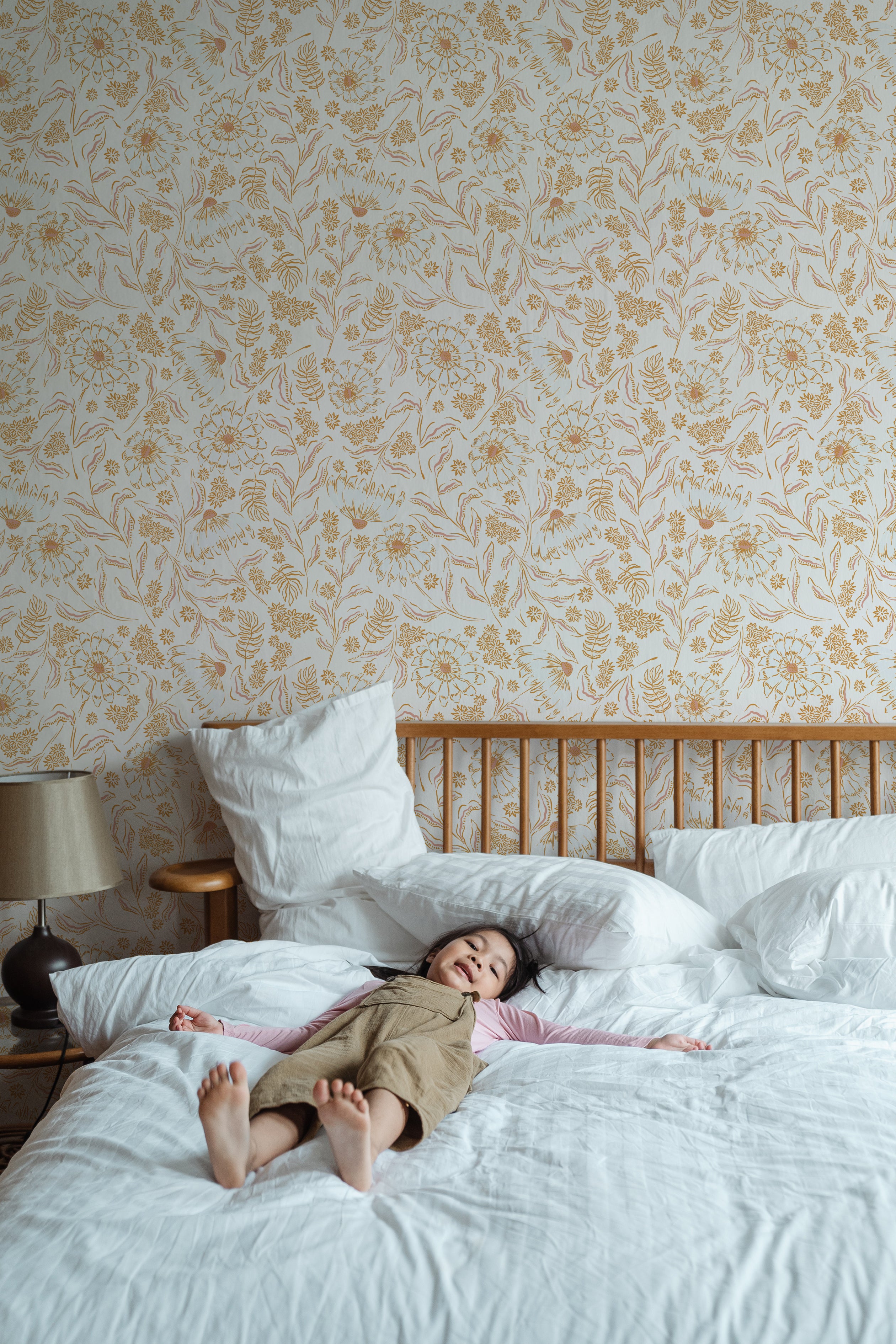 A young child lies relaxed on a large bed in a bedroom adorned with De Pijp Floral Wallpaper. The wallpaper showcases a soft golden floral design on a cream background, creating a serene and cheerful atmosphere. The room is furnished with a wooden bed and a vintage bedside lamp.
