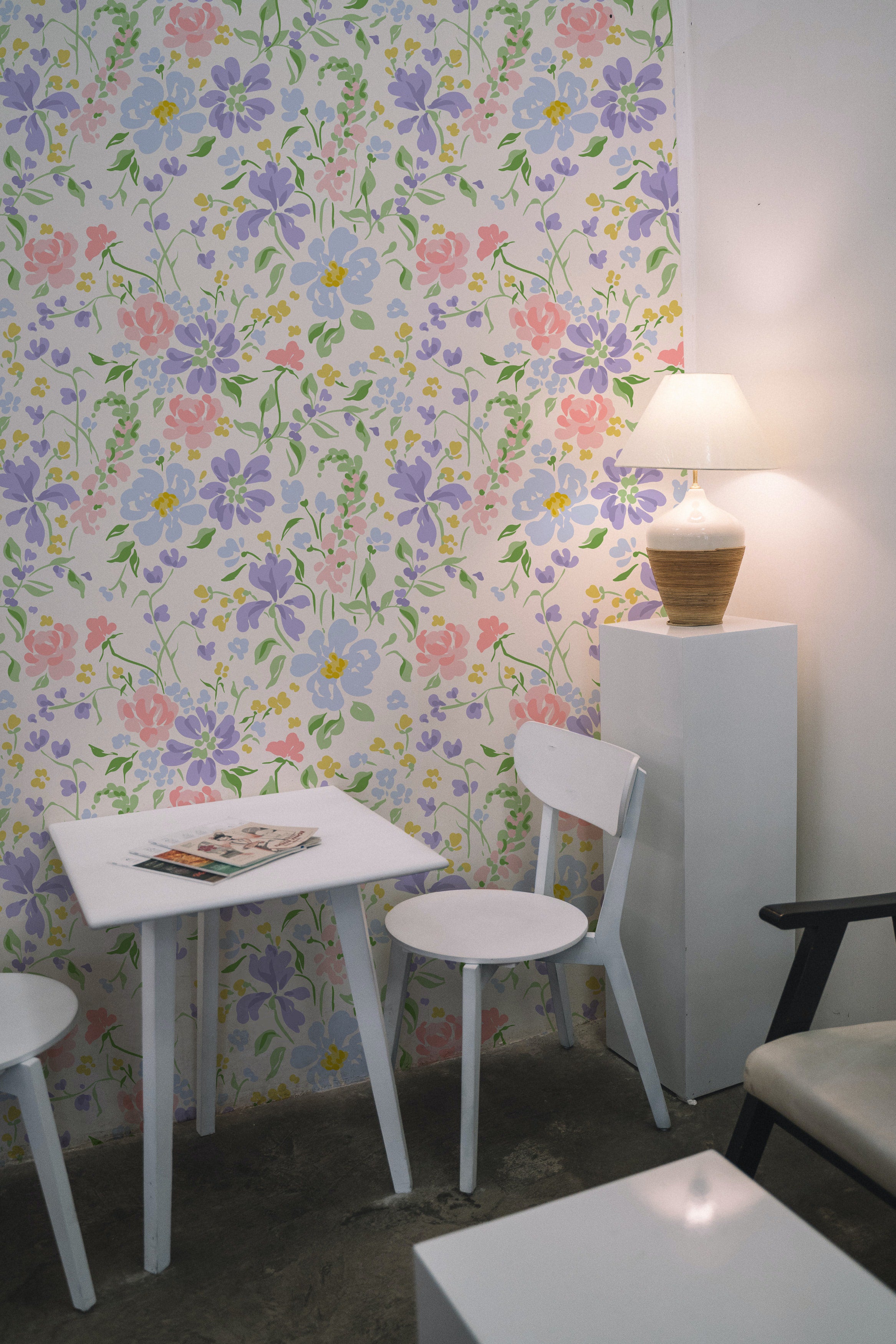 A simple yet stylish workspace with Romana Wallpaper, adorned with an array of colorful flowers in pink, blue, and yellow. A white table and chairs set against the floral backdrop creates a bright and airy study area