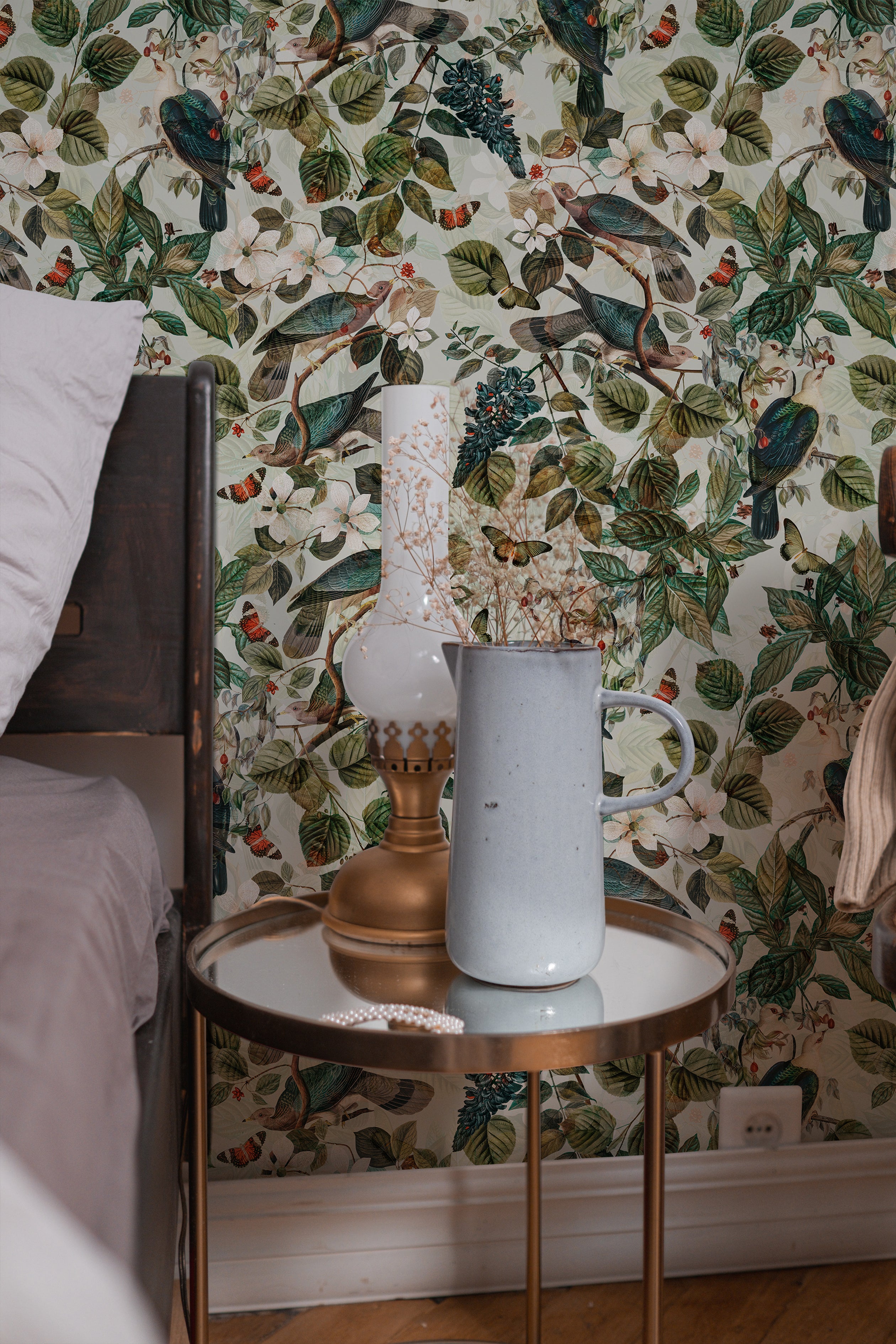 Charming bedroom corner decorated with Spring Pigeon Wallpaper illustrating a detailed pattern of green foliage, white flowers, and colorful pigeons. A bedside table holds a lamp and a mug, complementing the naturalistic theme.