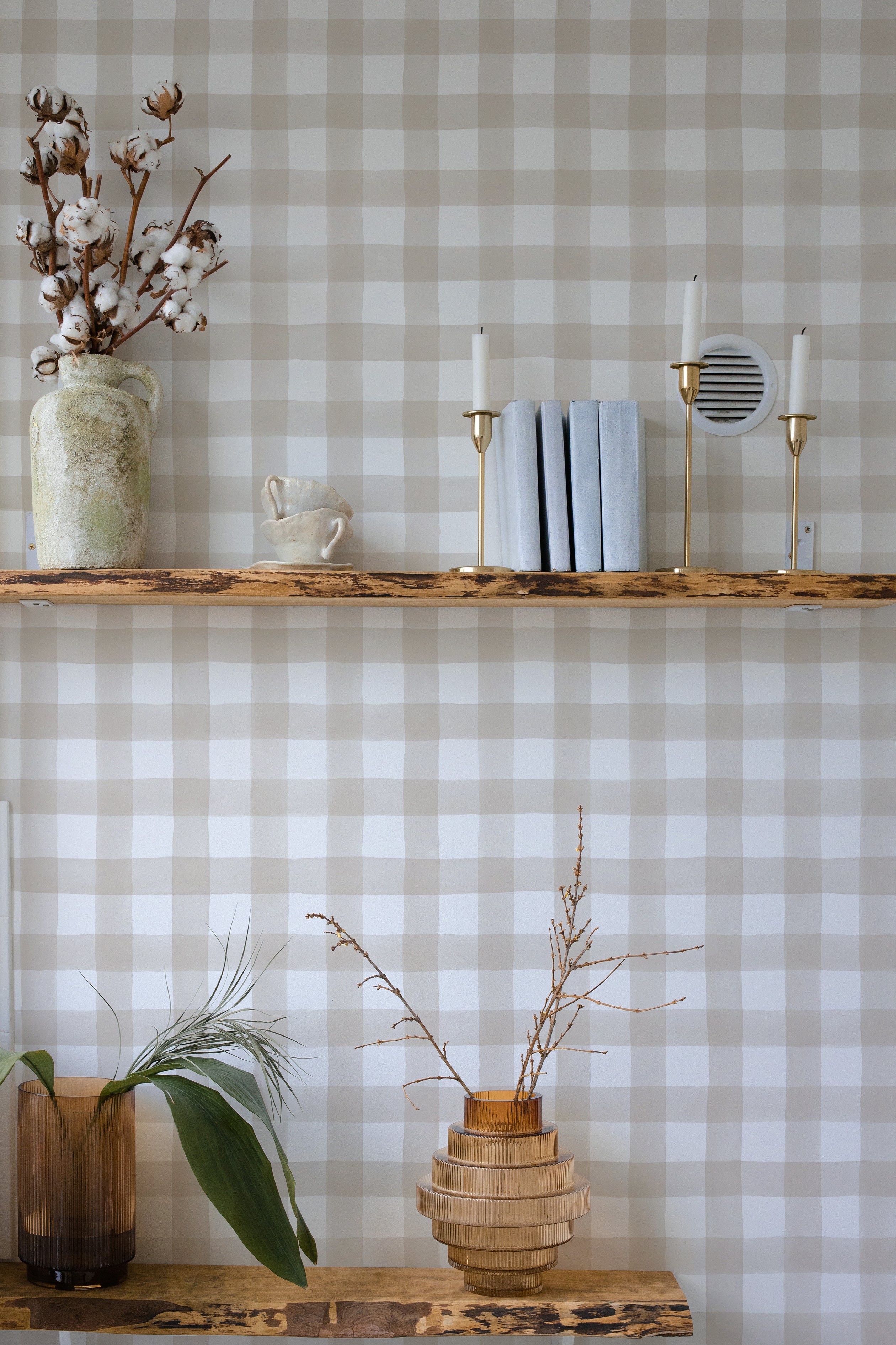 A rustic home decor scene with a wall adorned in beige and white buffalo check wallpaper. The scene includes a wooden shelf with a vase holding cotton branches, a ceramic cup, and modern candle holders, providing a warm and natural atmosphere.