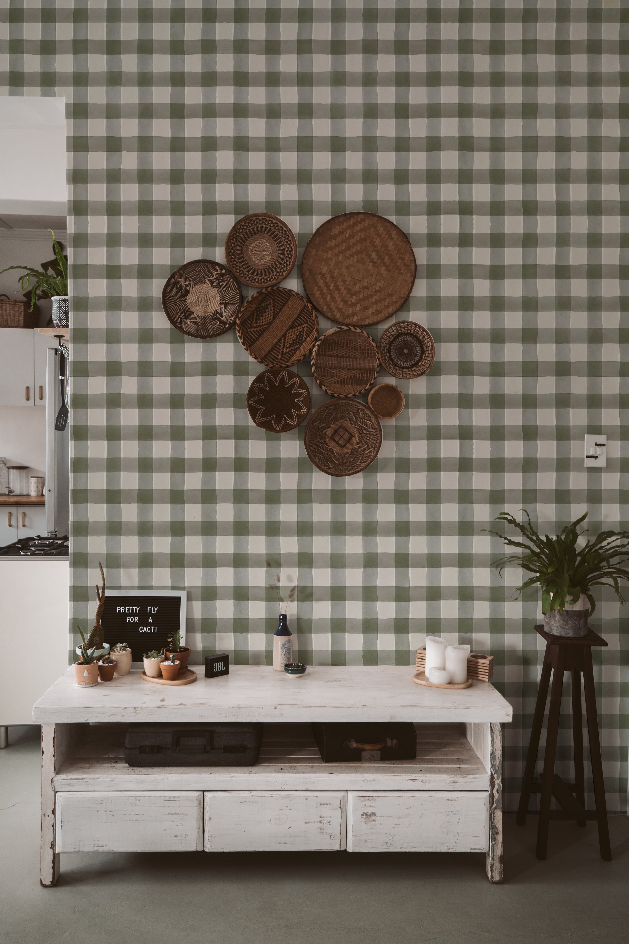 A contemporary kitchen scene with a wall adorned in Cuadro de Búfalo wallpaper featuring a refreshing green and white buffalo check pattern. The scene is decorated with a rustic white table, assorted woven baskets, and small potted plants, creating a vibrant and homey atmosphere.