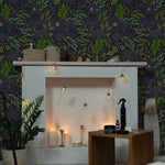 A cozy, warmly lit living area featuring the Martinique Floral Wallpaper, characterized by its large-scale dark purple flowers and lush green leaves on a dark background. The wallpaper creates a bold statement while providing a backdrop to a serene space decorated with candles and string lights.