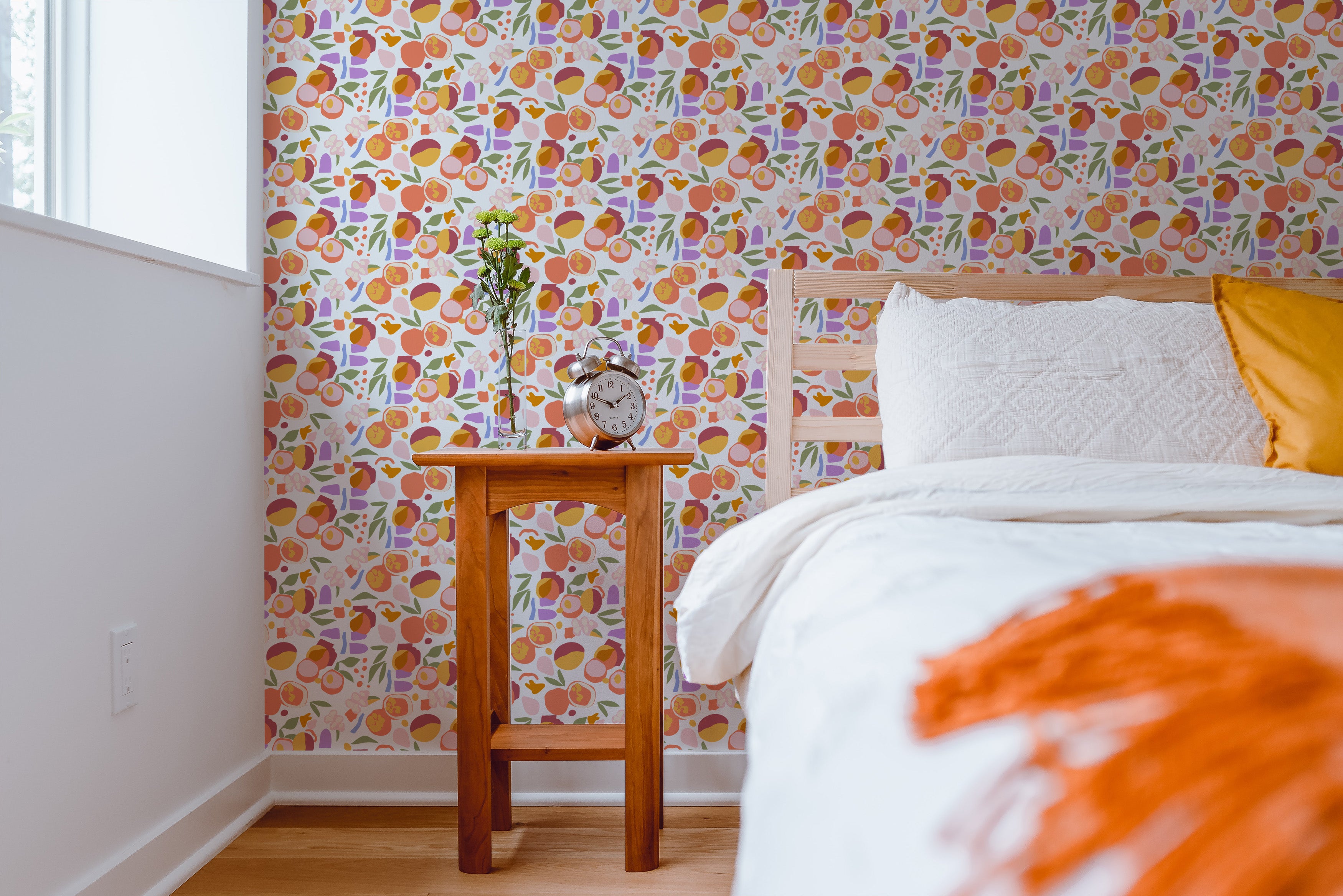 A bedroom scene showcasing the Funky Fruit Wallpaper with a vibrant array of fruit illustrations and floral elements on the walls. A white bed with orange and yellow pillows complements the lively wallpaper, enhancing a modern and youthful atmosphere in the room.