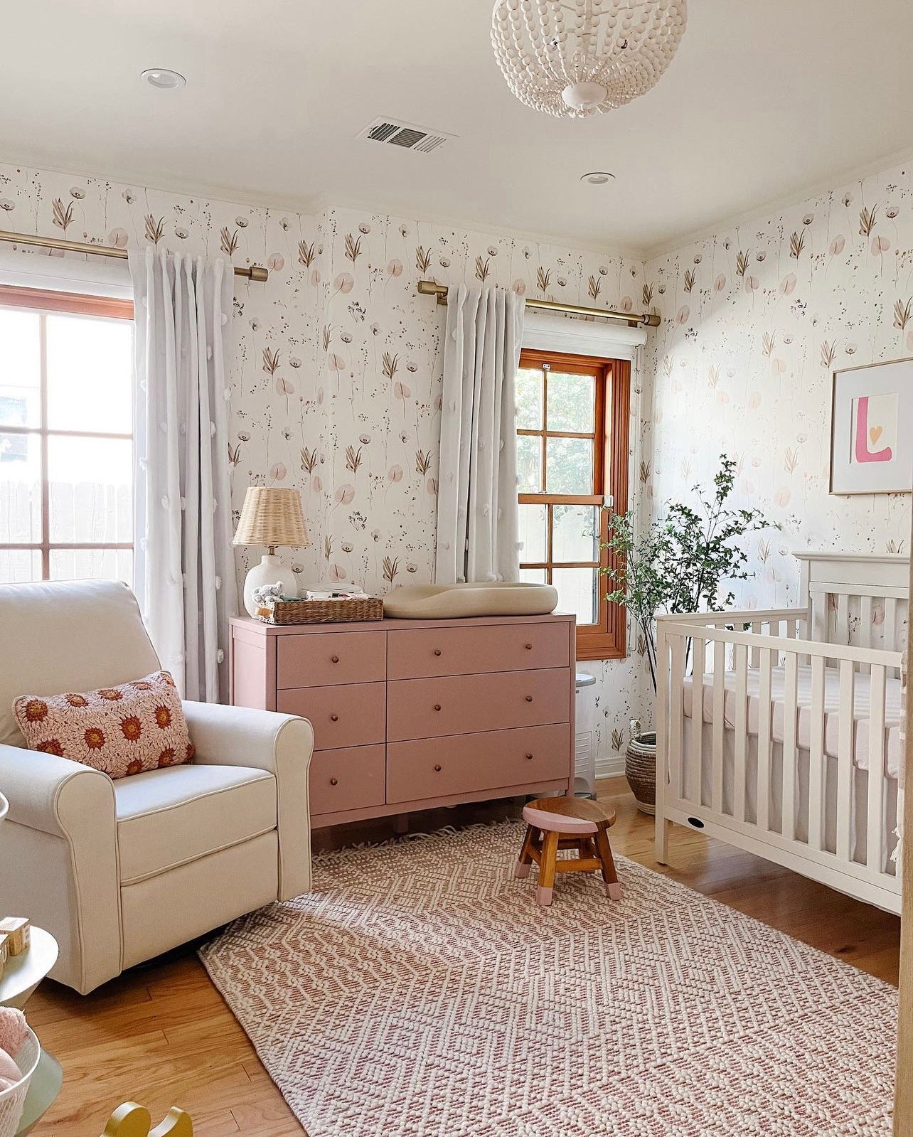 A nursery room featuring Ikebana Floral Wallpaper with a pattern of pastel flowers and foliage, complemented by white curtains, a blush pink dresser, a cozy armchair, and a wooden crib, creating a soft and inviting atmosphere.