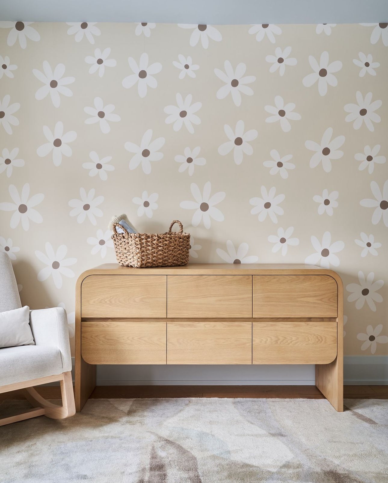 A modern home setting showing a wooden credenza against a wall covered with Simple Daisy Wallpaper. Above the credenza, a light-colored armchair and a woven basket enhance the room's modern yet cozy appeal.