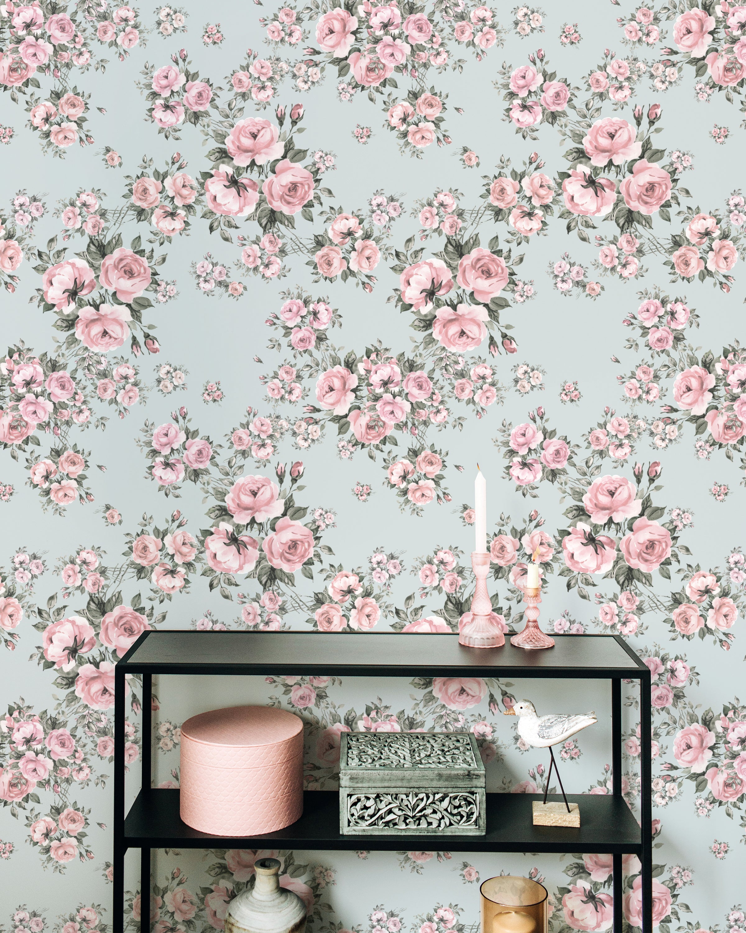 An elegant room with Rose Bouquet Wallpaper II showcasing a pattern of pink roses and green leaves on a light blue background. The decor includes a black shelving unit with candles, decorative boxes, and a bird ornament.