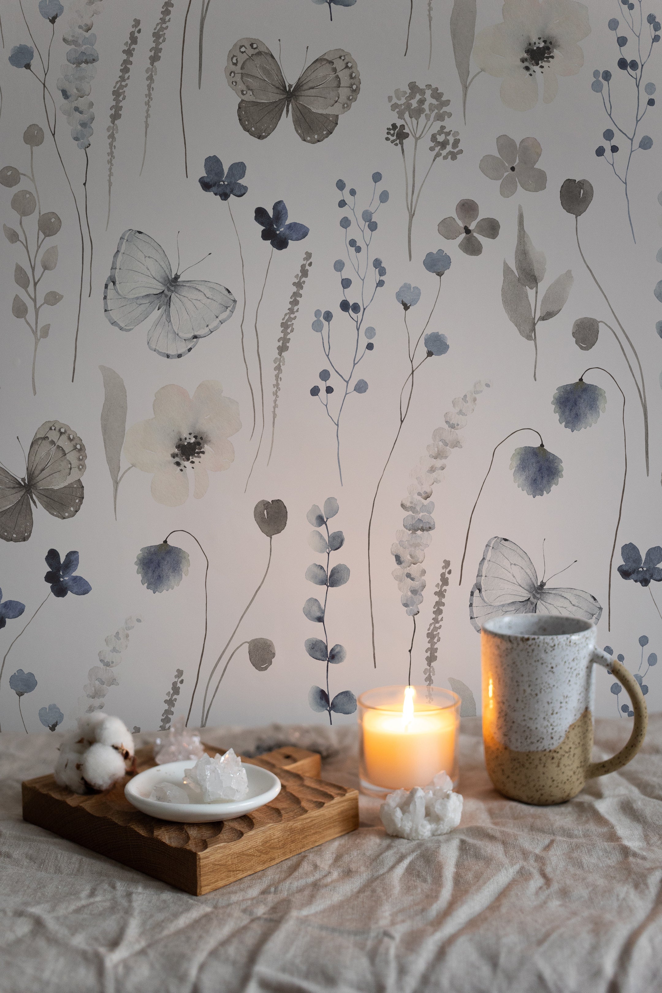 Romantic and warm atmosphere highlighted by Soft Flutter Wallpaper in a dimly lit setting with candles. The wallpaper's soft botanical patterns and butterflies provide a tranquil backdrop to a cozy setting with a mug and candles on a wooden tray