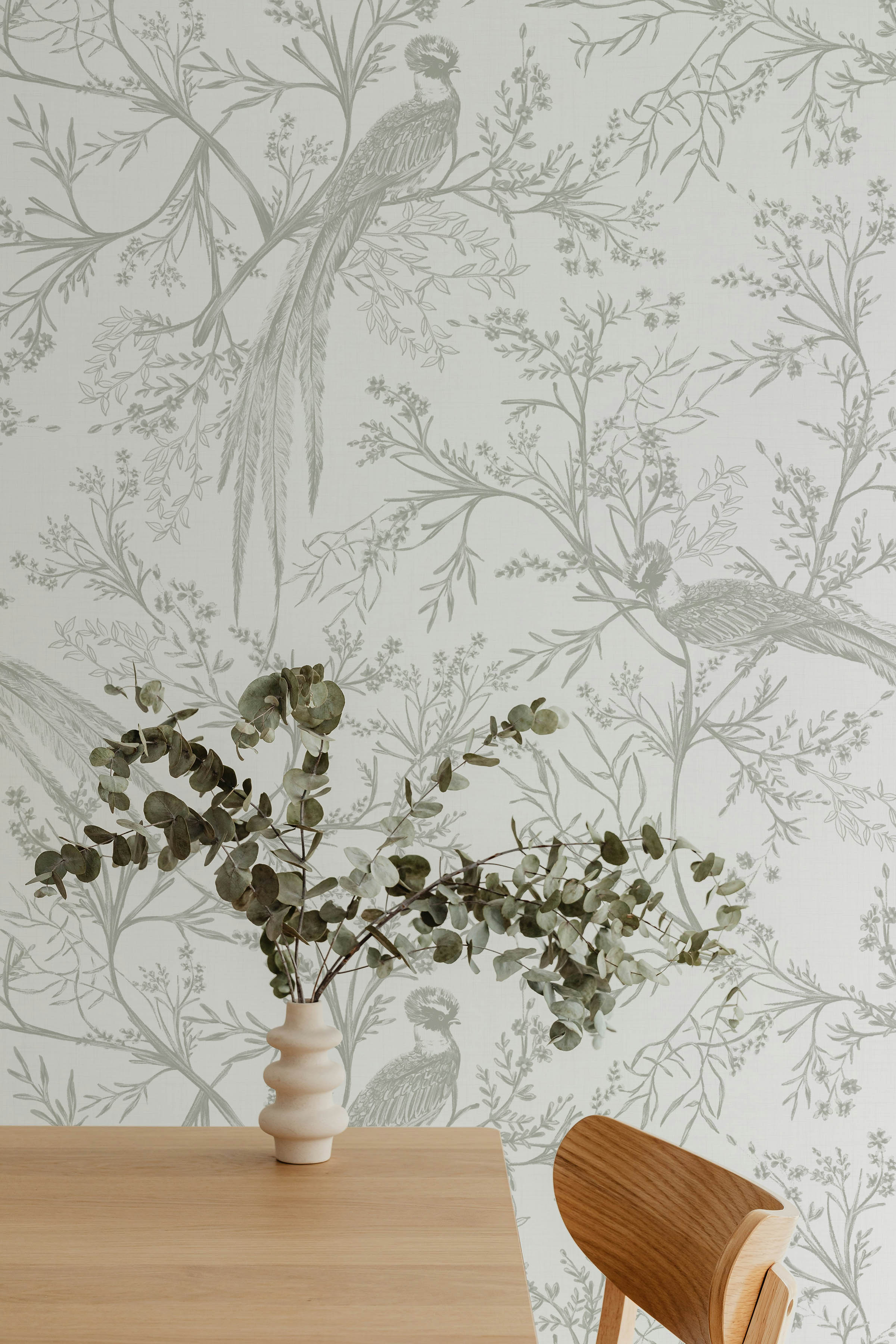 A minimalist style decor featuring the Oriental Garden Wallpaper, which beautifully complements a modern wooden table and a vase with silver dollar eucalyptus. The soft gray botanical prints against a white background add a touch of sophistication and calm to the space.
