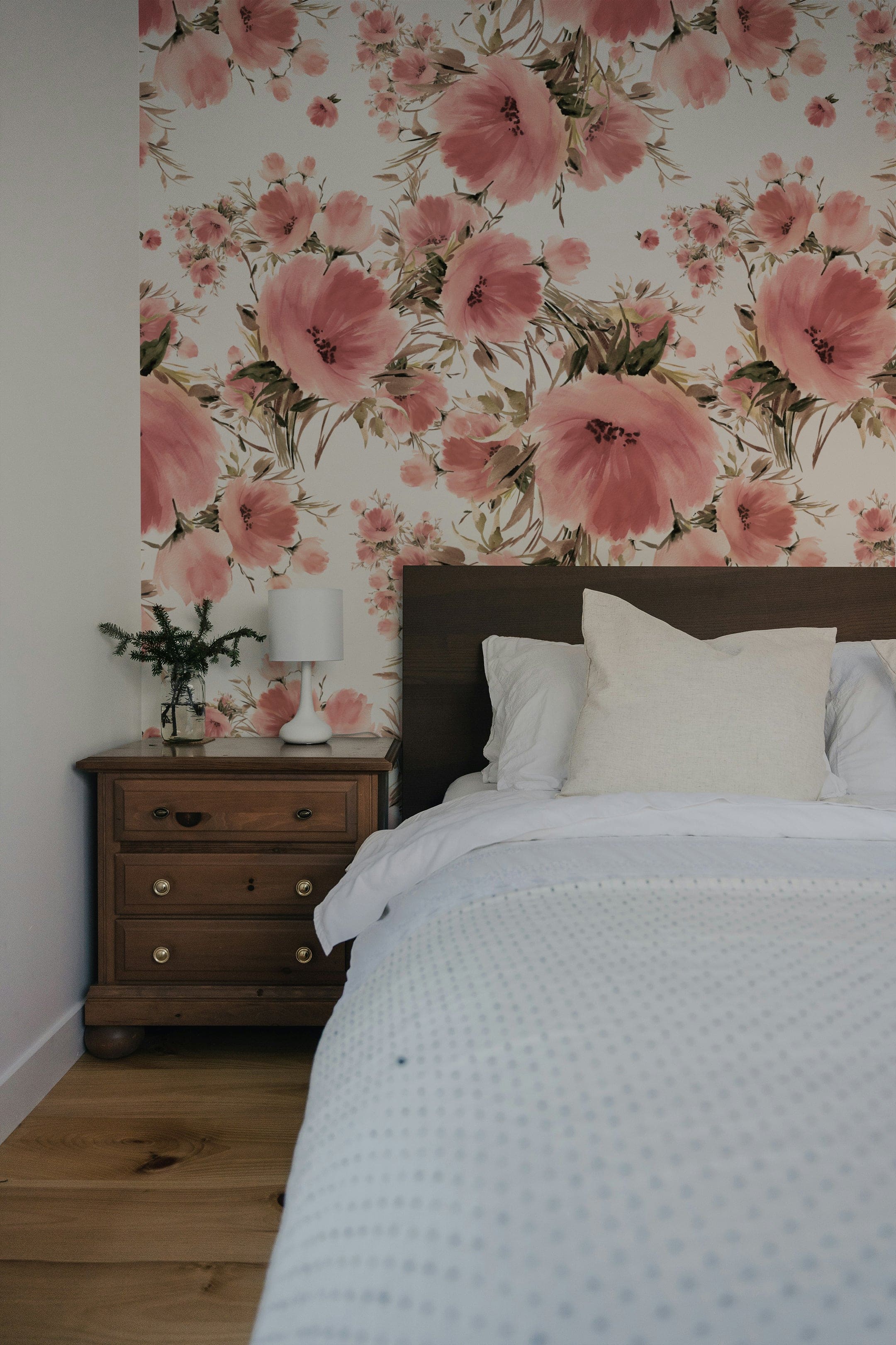 A bedroom interior showcasing one wall adorned with 'Grace Floral Wallpaper', which features large pink flowers and soft green leaves. The room includes a wooden bed with white bedding, a small wooden nightstand with a white lamp and a small pine tree decoration, creating a cozy and inviting atmosphere