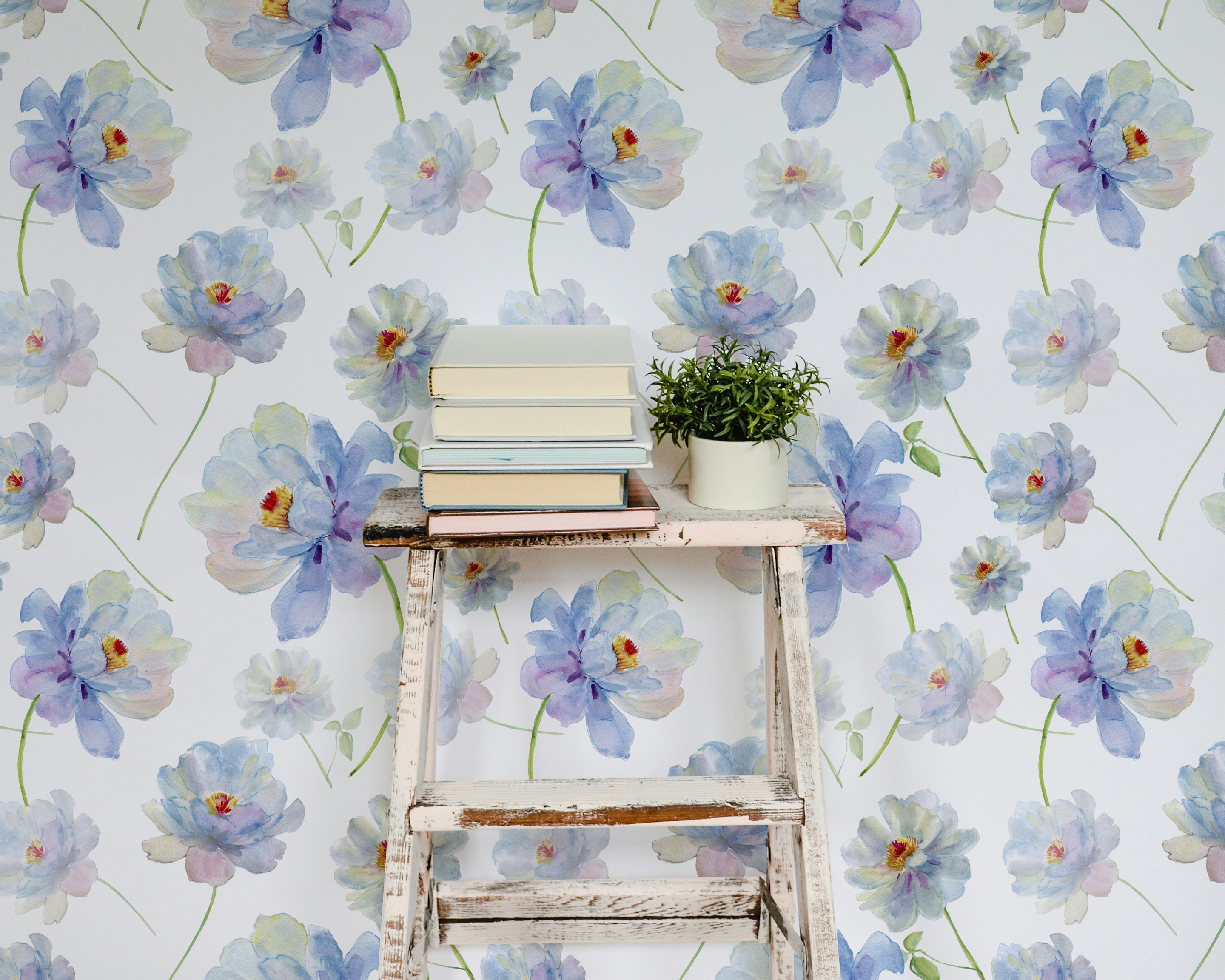 Cozy reading nook decorated with Watercolour Bliss Wallpaper, featuring large watercolor flowers. The setting includes an old white wooden ladder used as a shelf holding books and a small potted plant, adding a rustic charm to the floral decor