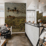 Vintage-style cafe interior with a rustic landscape wall mural. The scene on the wall creates a tranquil backdrop for the cafe setting, featuring wooden shelves with potted plants and simple seating arrangements