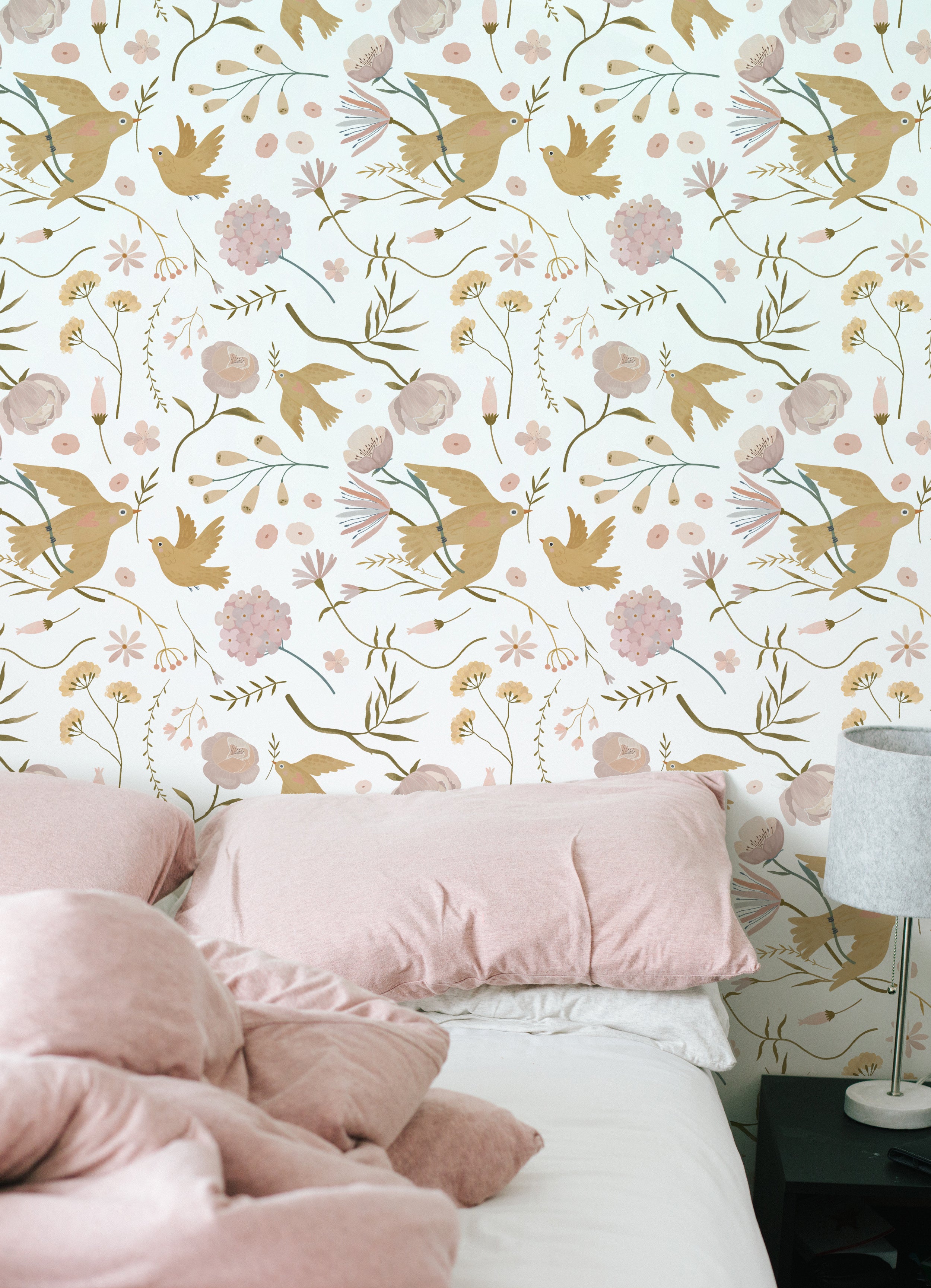 A cozy bedroom with the Pastel Garden Wallpaper creating a soothing backdrop. The wall is adorned with depictions of birds and delicate flowers in soft pinks and greens, complementing the plush pink bedding
