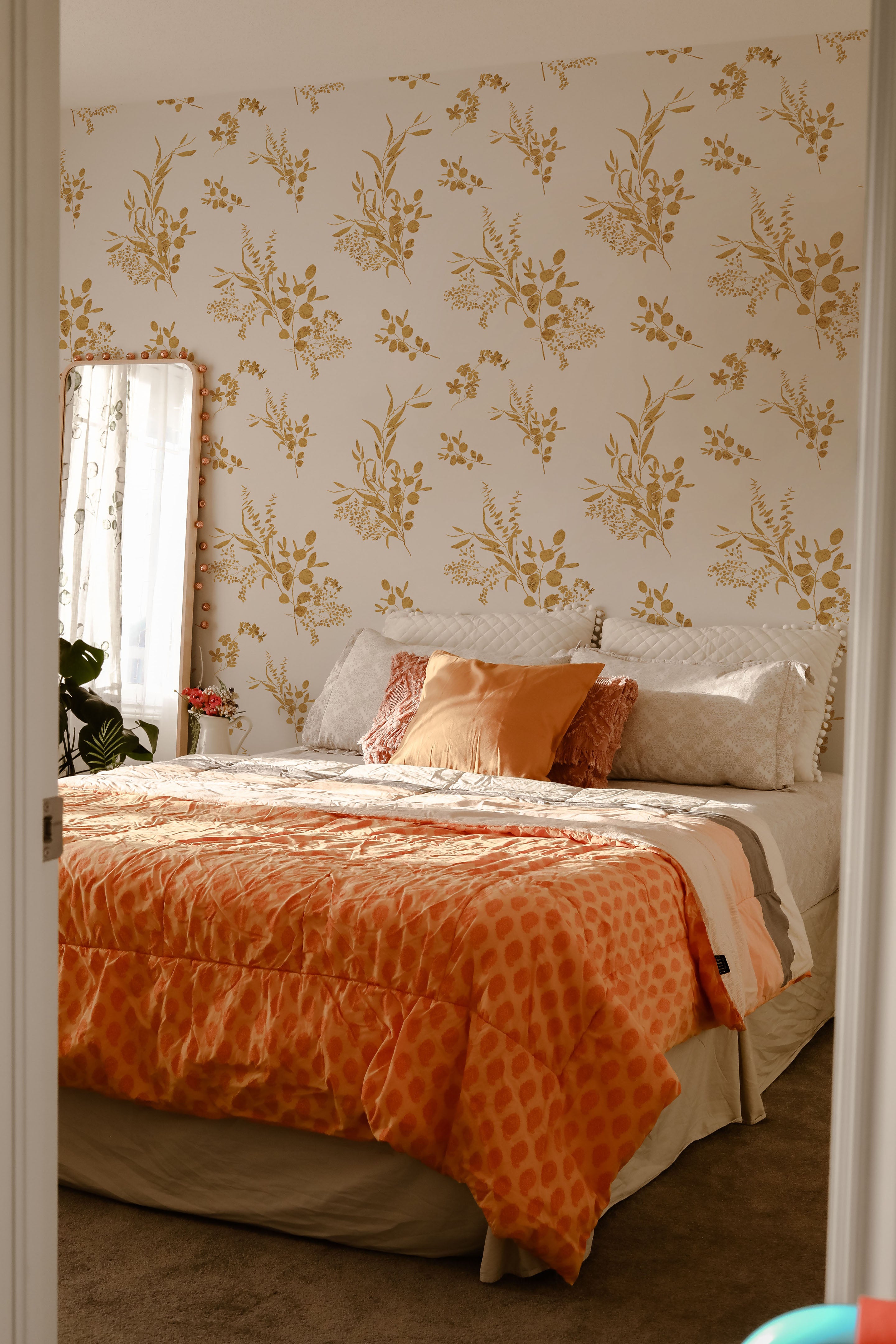 A serene bedroom showcasing the Golden Greenery Wallpaper, which adds a touch of nature-inspired elegance. The room includes a bed covered in a vibrant orange and beige patterned duvet, complemented by decorative pillows and a view into a sunlit window with sheer curtains.