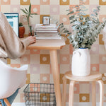 A home office setup enhanced by Coralie Wallpaper, showcasing a colorful grid pattern with peach, beige, and gold tones. The workspace is organized with a small desk, books, and a large potted plant, offering a calm and creative environment.