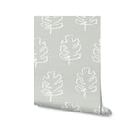A roll of Palm Leaves Wallpaper, displaying a serene pattern of white palm leaf outlines on a muted grey background, perfect for creating a peaceful and minimalist environment in any room.