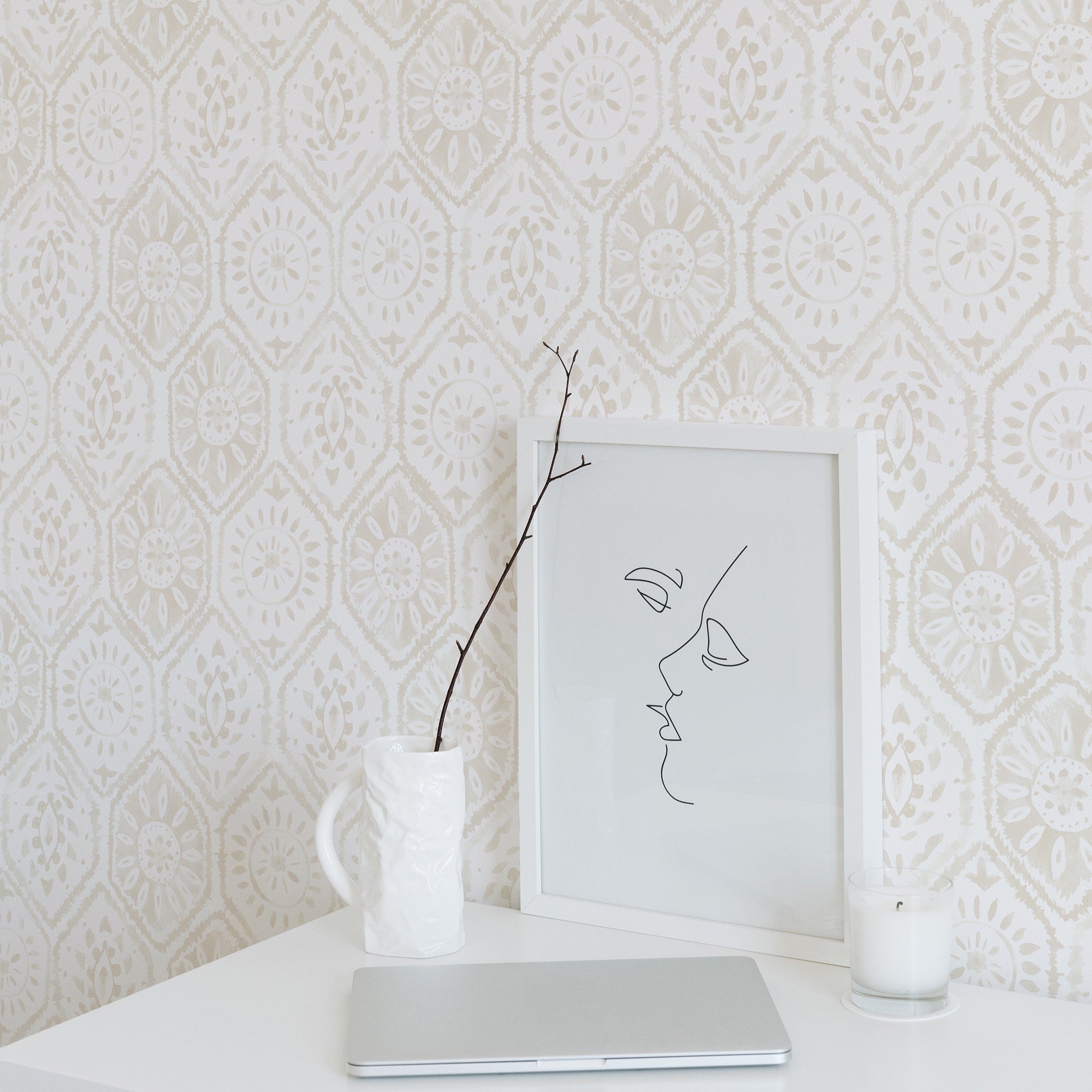 A minimalist workspace highlighted by the Moroccan Tile Wallpaper in Ecru. The wallpaper's intricate pattern provides a soft, decorative background that pairs well with the simple desk, modern line art in a frame, a white laptop, and a single branch in a vase, creating a serene and stylish area.
