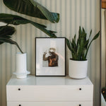  A serene bedroom setup featuring a white dresser topped with a framed black and white photo of a playful moment at the beach. A large green plant in a white pot sits beside the frame, enhancing the natural ambiance. The room is characterized by soft, striped green wallpaper, adding a subtle texture to the calm, well-organized space.