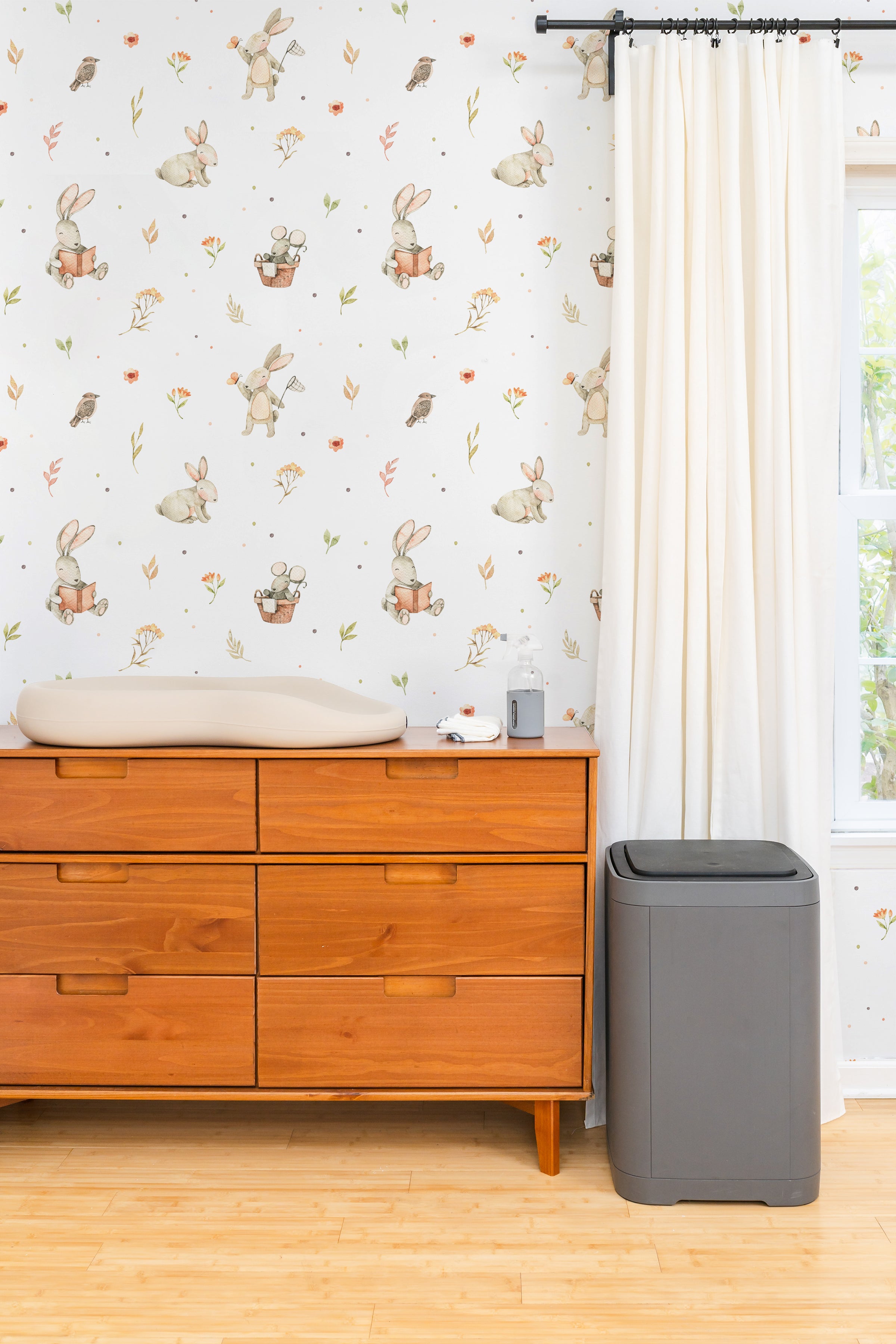 A nursery room adorned with Watercolour Bunnies Wallpaper creating a gentle and whimsical ambiance. The wallpaper showcases soft-toned bunnies, birds, and flora, complementing the room's calm decor and wooden furniture