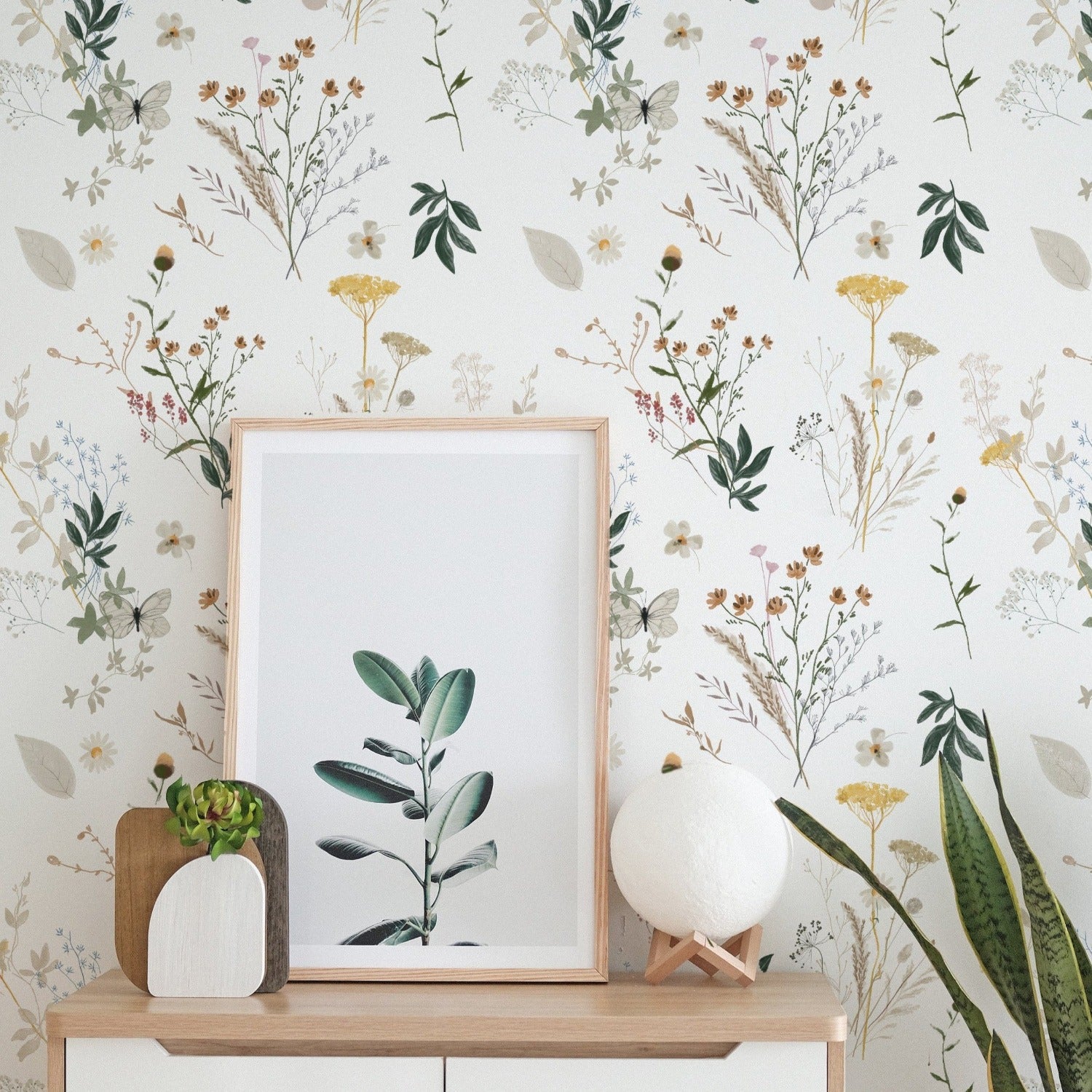 A wall adorned with floral wallpaper with a delicate illustration of wildflowers and leaves in a muted color scheme. A modern Scandinavian-style sideboard displays a framed picture of green foliage, a small white vase, and a textured spherical decor item.