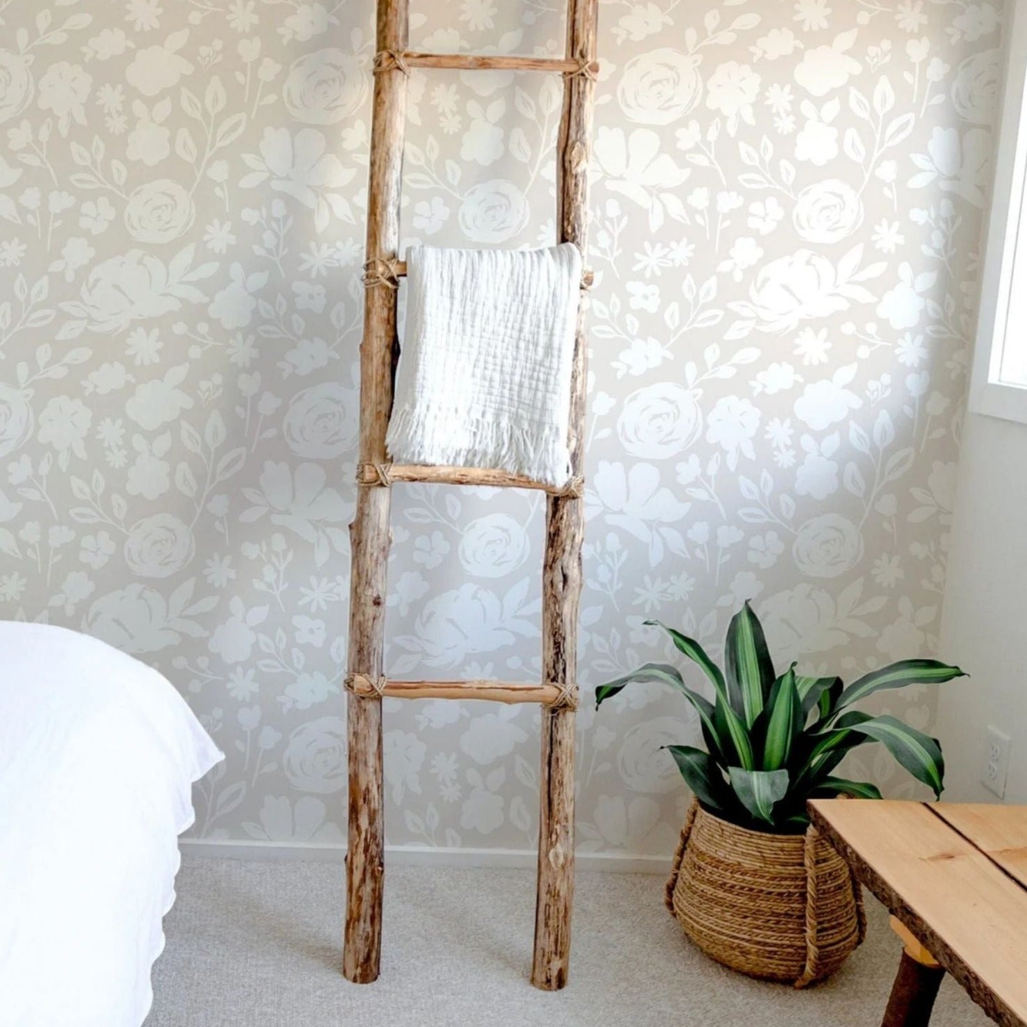 A bedroom setting showcases the Silhouette Floral Wallpaper, creating a tranquil ambiance with its faded linen floral silhouettes. A rustic wooden ladder serves as a unique decorative piece, draped with a white towel, next to a potted plant, enhancing the room’s natural and calm aesthetic.