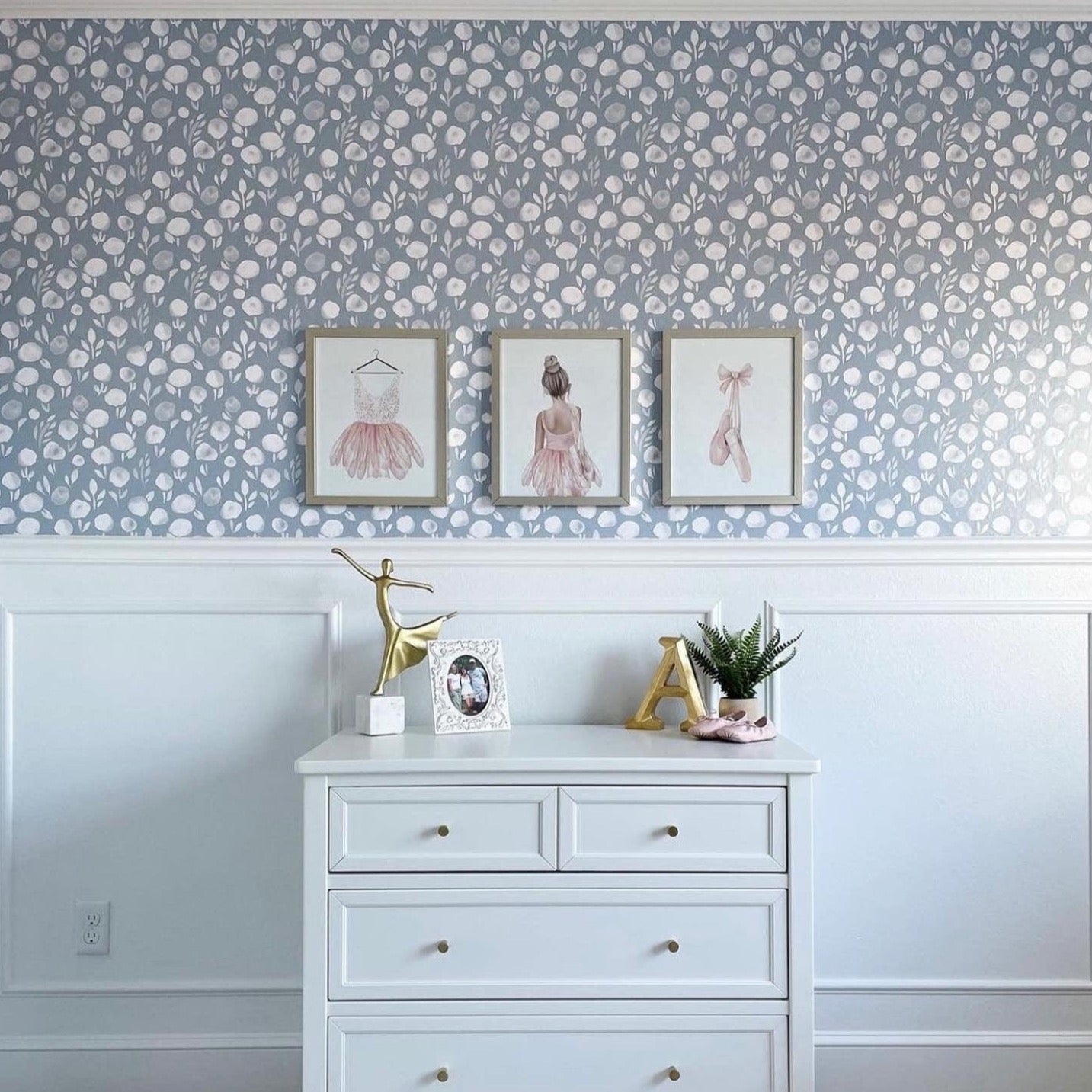 A tastefully decorated wall featuring the 'Subtle Botanica II - Inverted' wallpaper above wainscoting, accented with framed artwork of ballet attire and a chic dresser, creating an elegant space that is both sophisticated and whimsical.