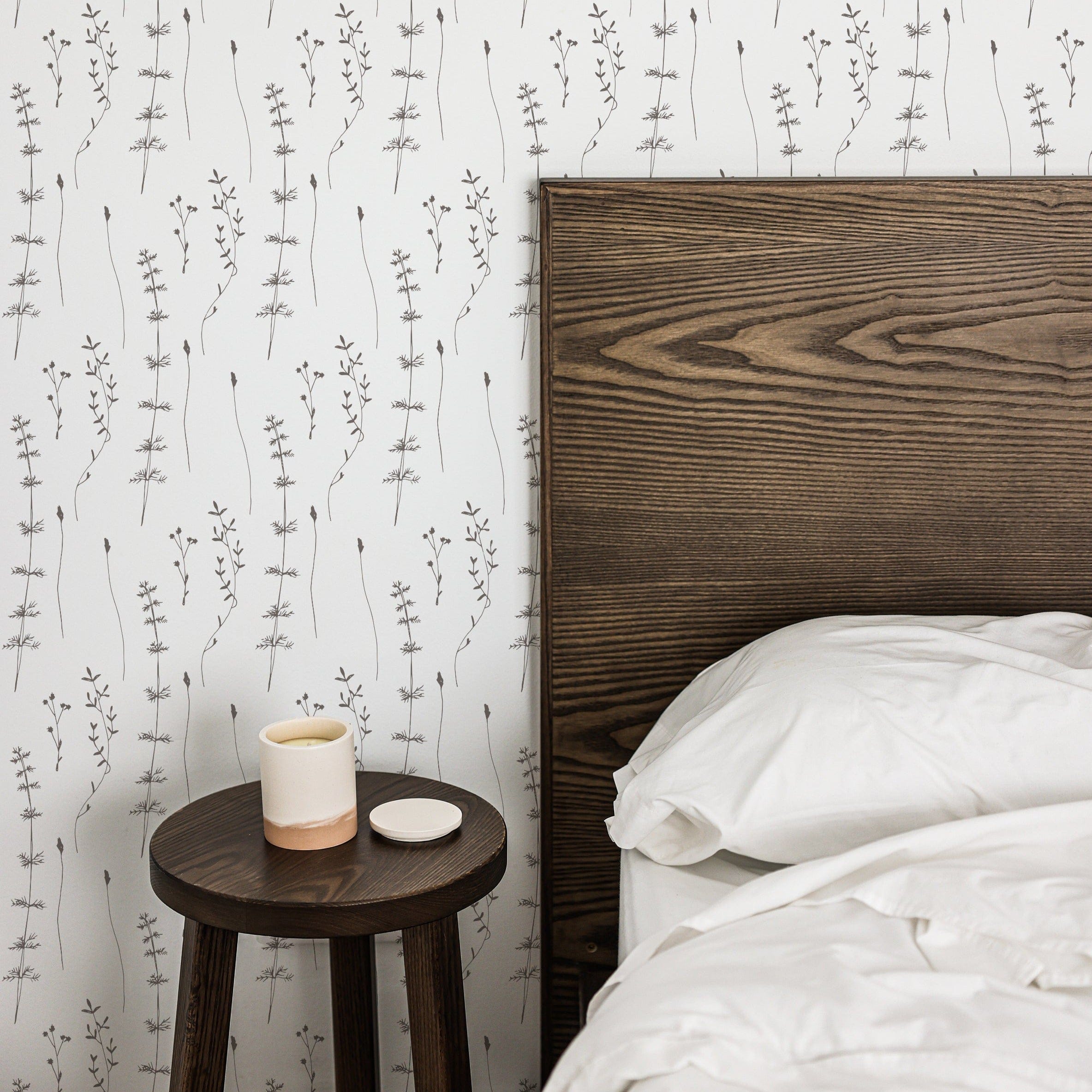 A minimalist bedroom scene featuring Delicate Wildflower Wallpaper. A wooden bed frame adjacent to a small round side table, which holds a ceramic candle holder, enhances the organic and tranquil ambiance created by the floral pattern.