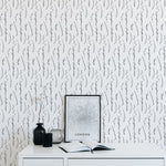 A minimalist home office setup featuring Twig & Leaf Wallpaper with a black and white pattern of delicate twigs and leaves, complemented by a white desk, a framed picture, and black decorative items.
