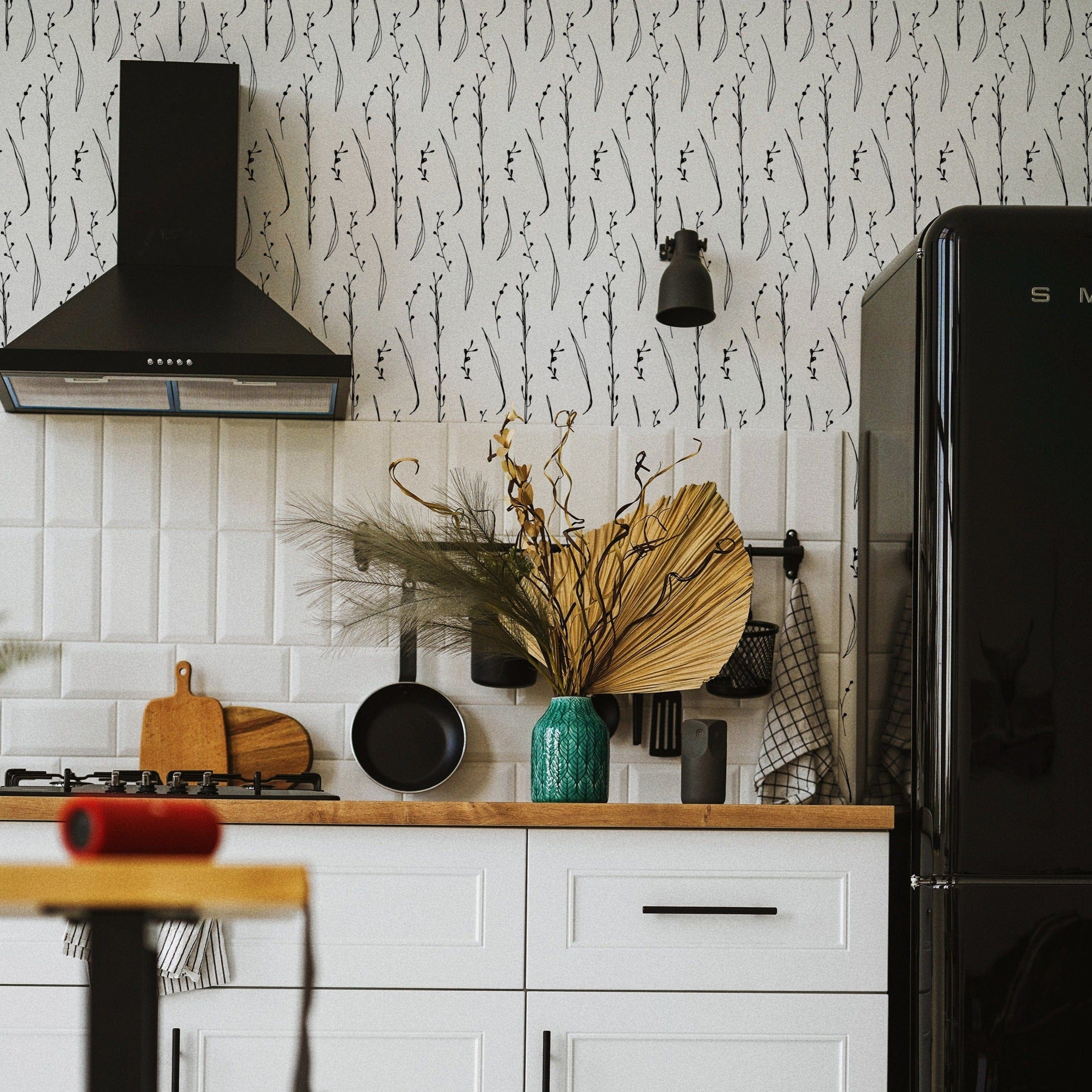 A modern kitchen featuring Twig & Leaf Wallpaper with a black and white pattern of twigs and leaves, adding a stylish touch to the white cabinets and black appliances.