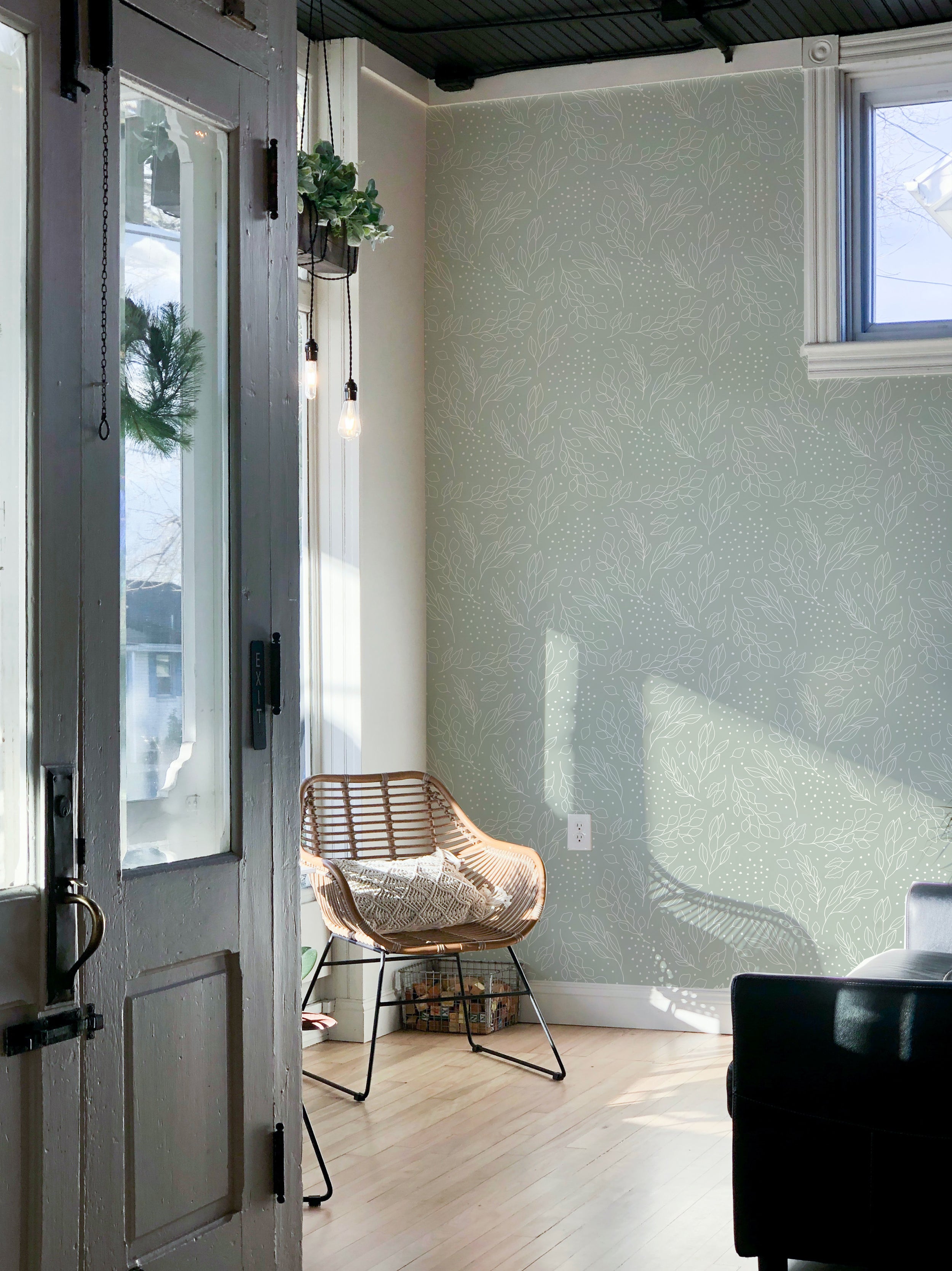 A tranquil entryway featuring Ferns and Leaves Wallpaper, showcasing a subtle leaf pattern in soft green tones. The decor includes a rattan chair by the window and vintage style hanging lights, creating a warm and welcoming atmosphere.