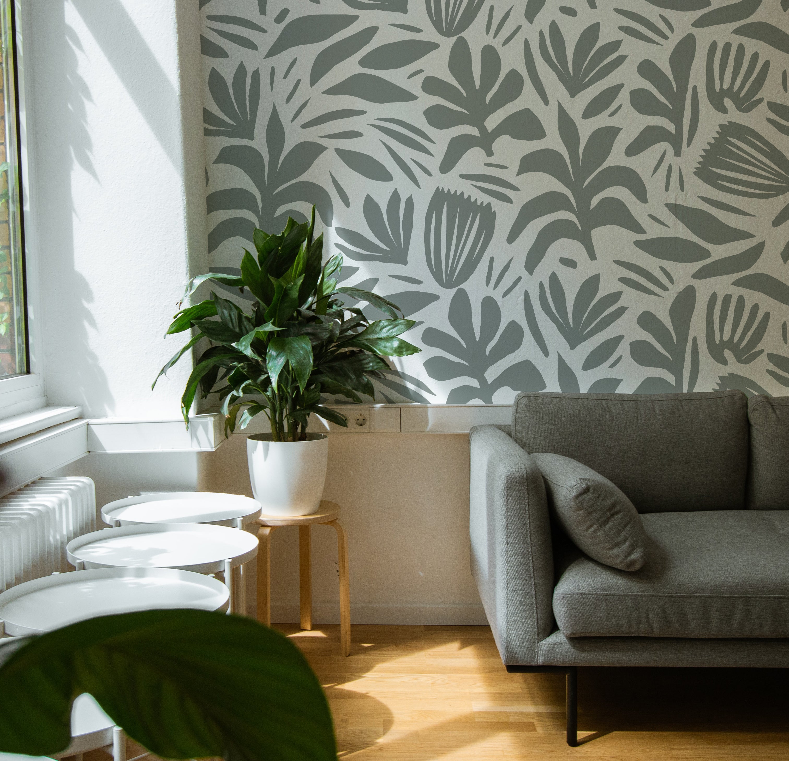 A modern living room enhanced by the Night Train Abstract Floral Wallpaper, adding a touch of elegance with its abstract floral design in gray tones. The room is complemented by a contemporary gray sofa and a vibrant green houseplant.