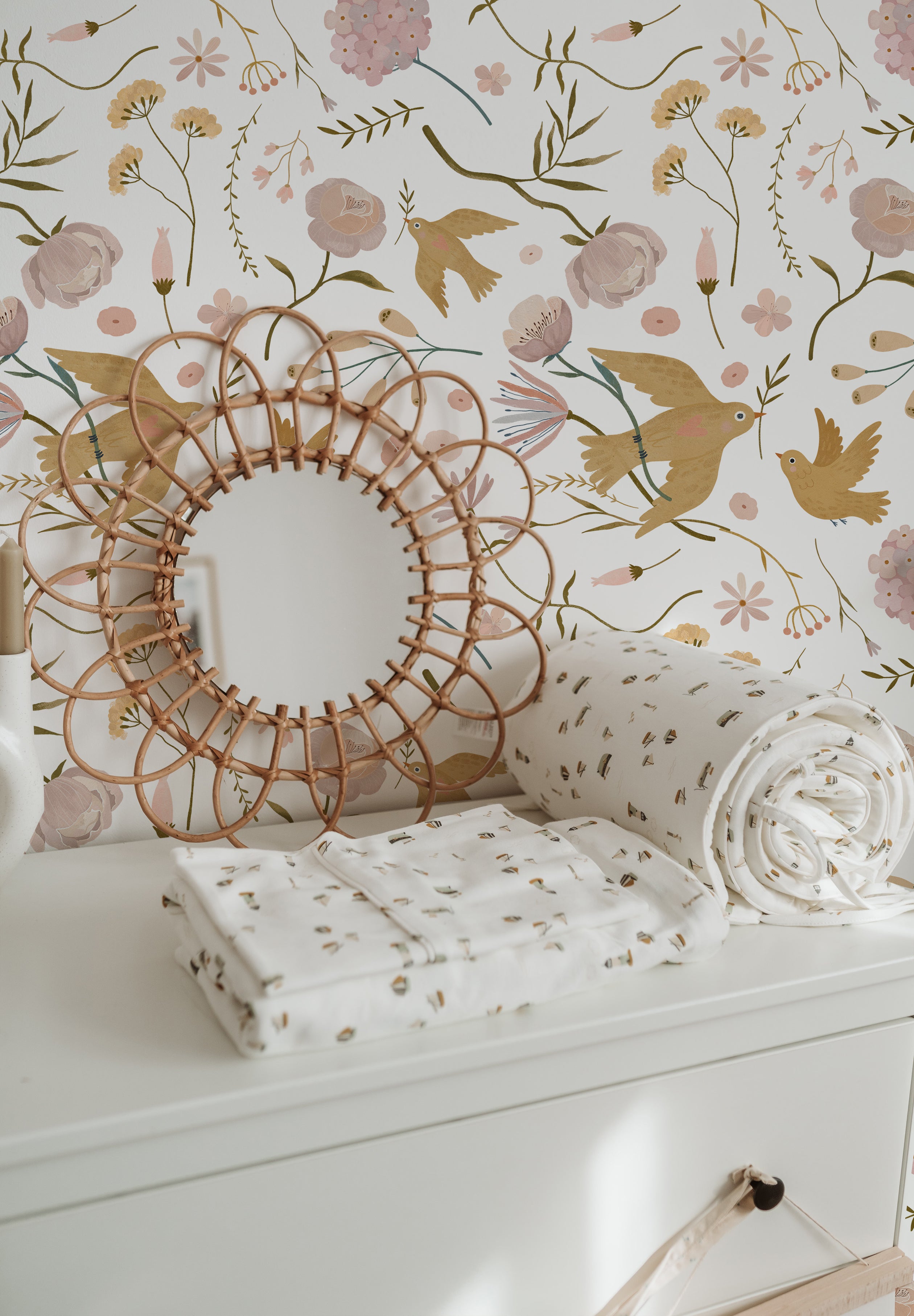 A decorative setting featuring the Pastel Garden Wallpaper, with a woven circular mirror and floral-printed textiles enhancing the wallpaper's intricate pattern of birds and blossoms.