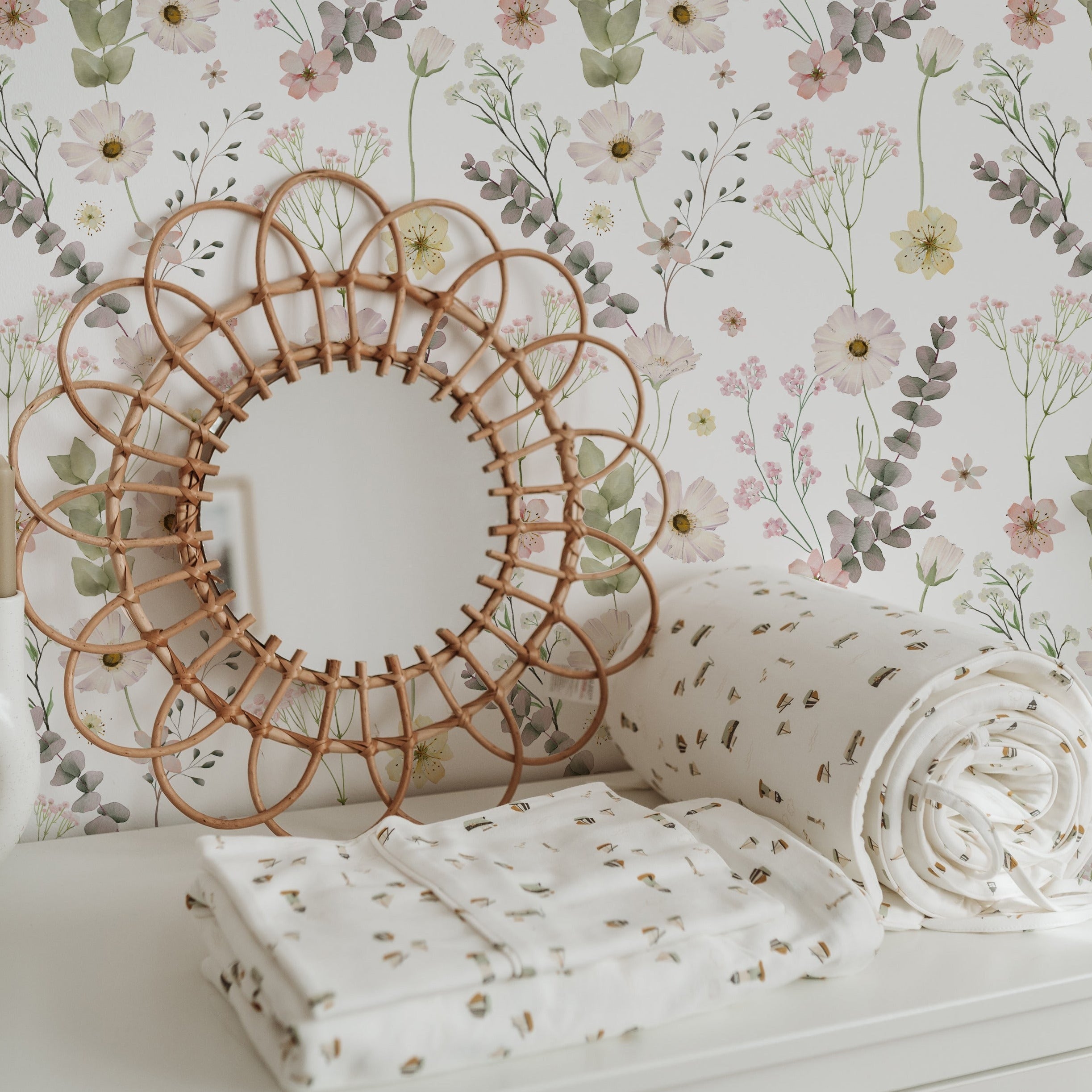 A close-up of a nursery room corner with a woven circular mirror above a white dresser. The Botanical Muse Wallpaper provides a soft backdrop with its floral design, complemented by a heart-shaped pillow and white bedding with a botanical print.