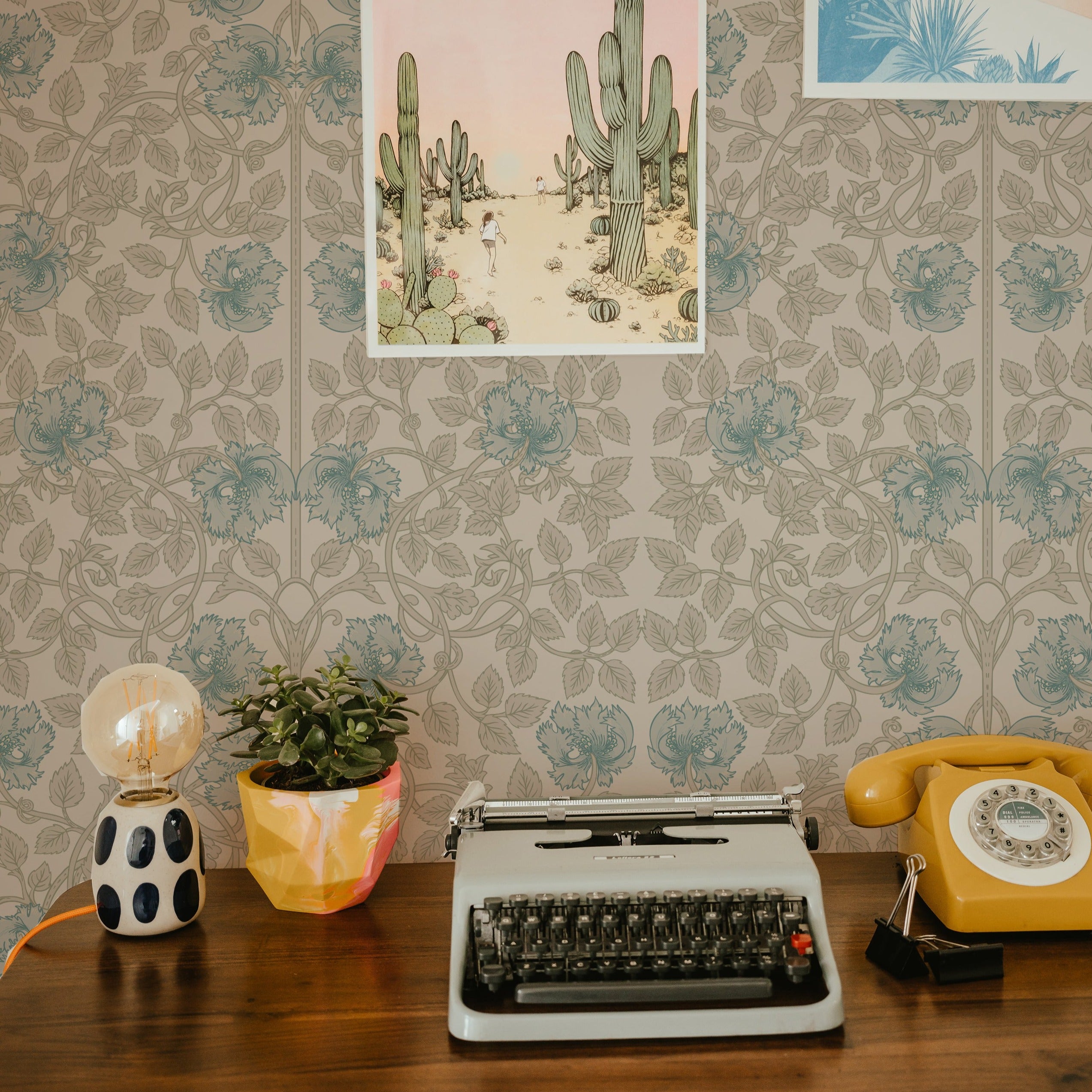 A vintage-inspired workspace with a retro typewriter, a rotary dial phone, and a modern table lamp on a desk. The background is adorned with a Color Burn Damask Wallpaper in teal and beige floral patterns