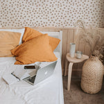 A cozy and inviting bedroom setup with a laptop open on a white bed, surrounded by mustard yellow pillows. The room features a floral wallpaper with small black and peach flowers, enhancing a warm, serene atmosphere.