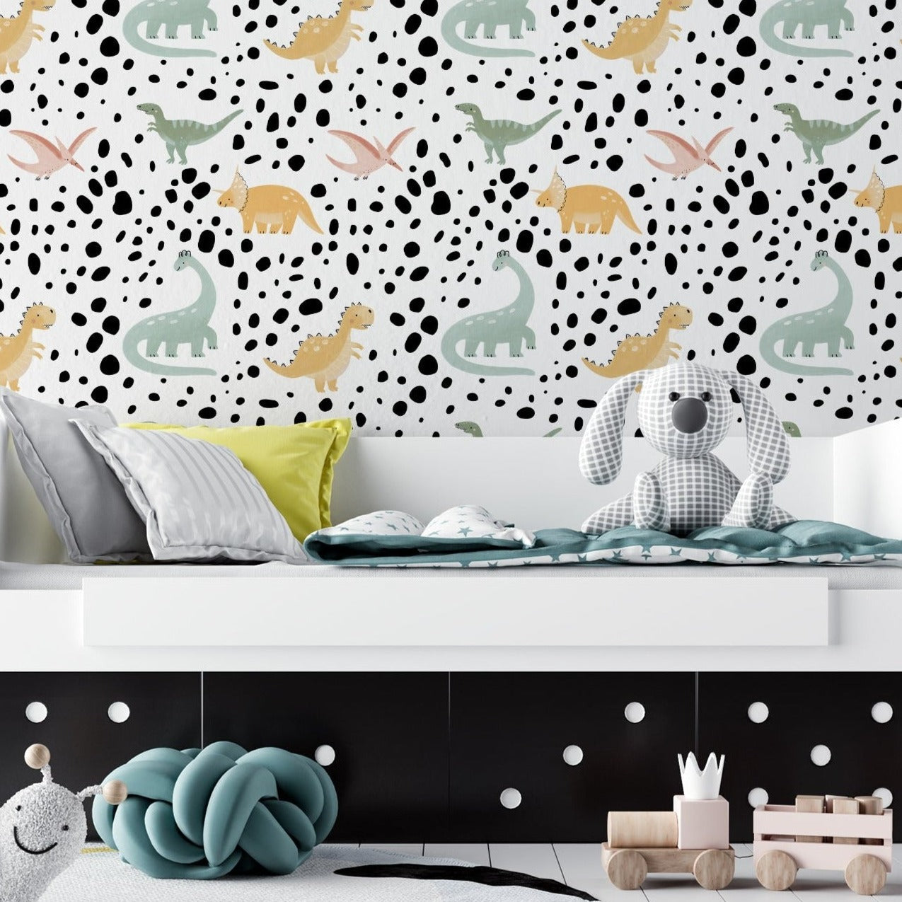 Children's room featuring a wall covered in 'Dino World Kids Wallpaper III' with pastel-colored dinosaur illustrations and black speckles on a white background. The room includes a grey bed with pillows, a plush grey dog toy, and various children's toys on the floor.