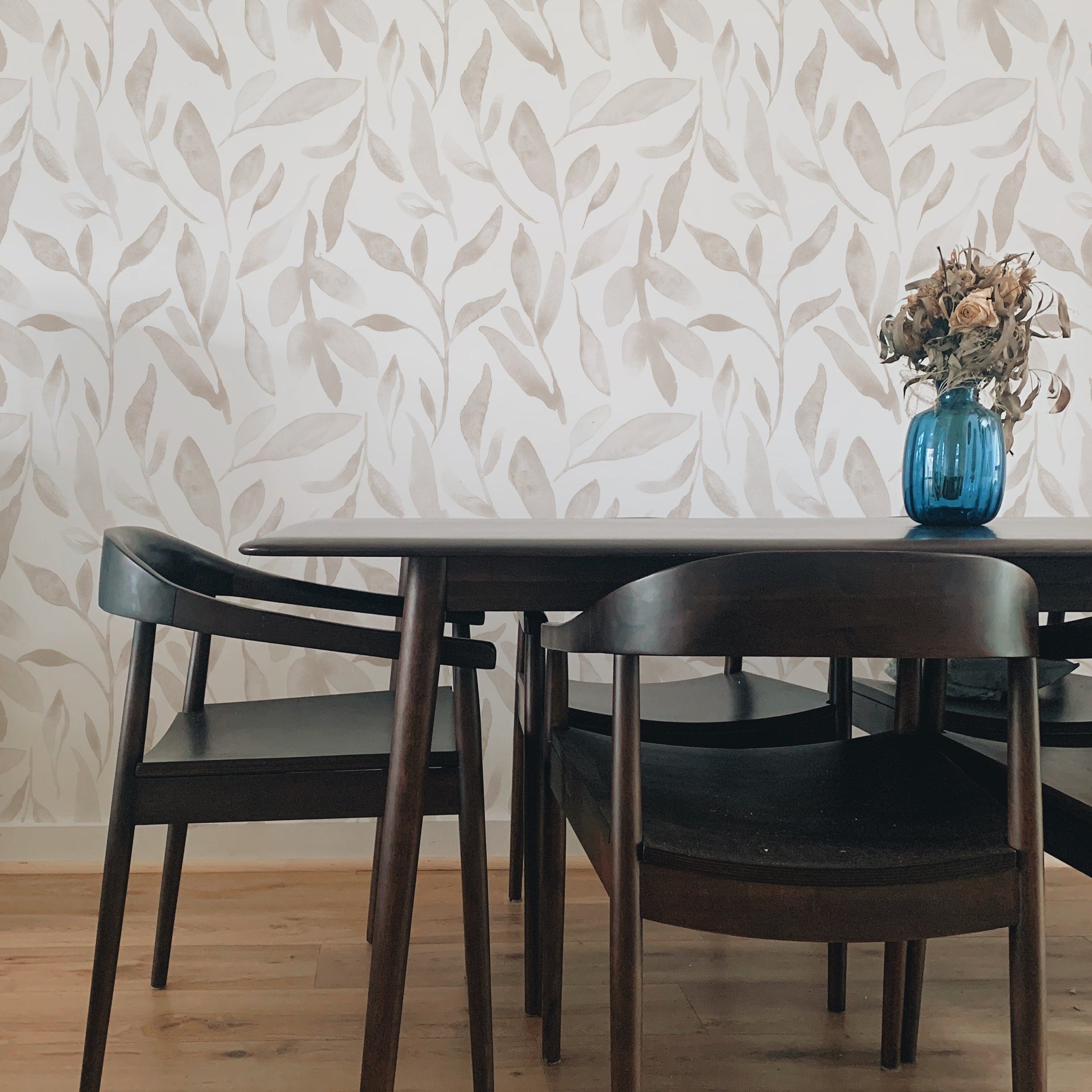 Contemporary dining area with Delicate Watercolour Leaves Wallpaper enhancing the space with its subtle beige leaf designs on a white background. The room features a dark wooden table, complemented by vintage-style chairs and a vibrant blue vase, merging modern aesthetics with classic elements.