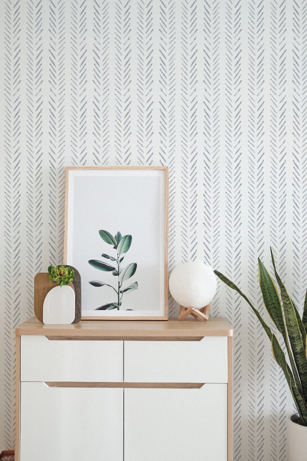 Minimalist and stylish room with Hand Painted Chevron Wallpaper, a framed botanical print, a white round lamp, and a wooden sideboard with decorative items.