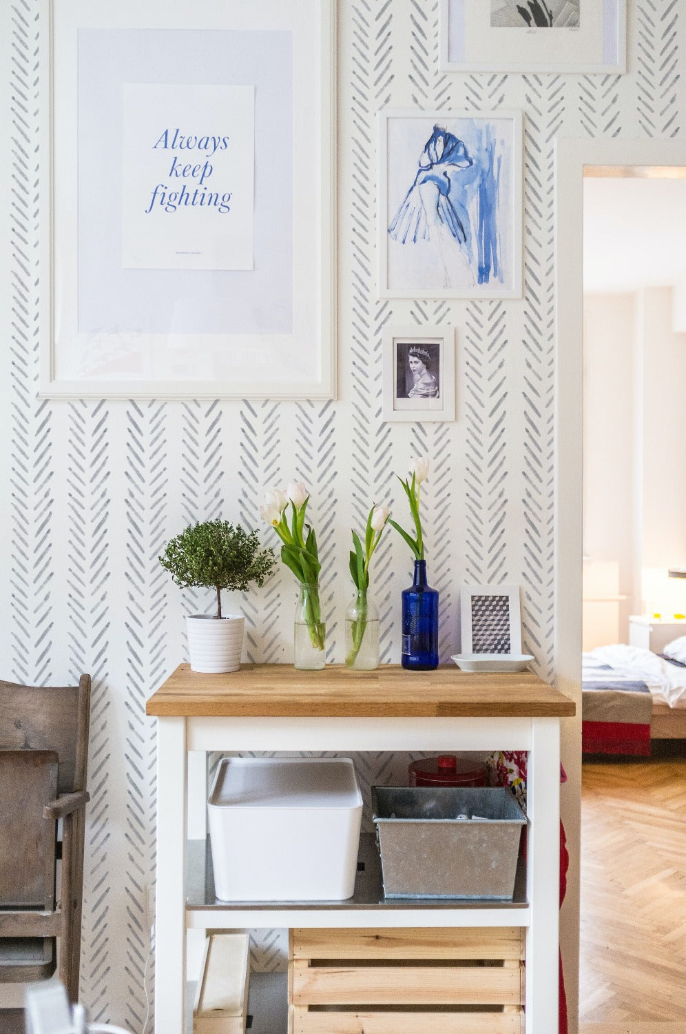 Eclectic workspace with Hand Painted Chevron Wallpaper, featuring a collection of framed prints, fresh flowers, and a wooden shelf with storage boxes.